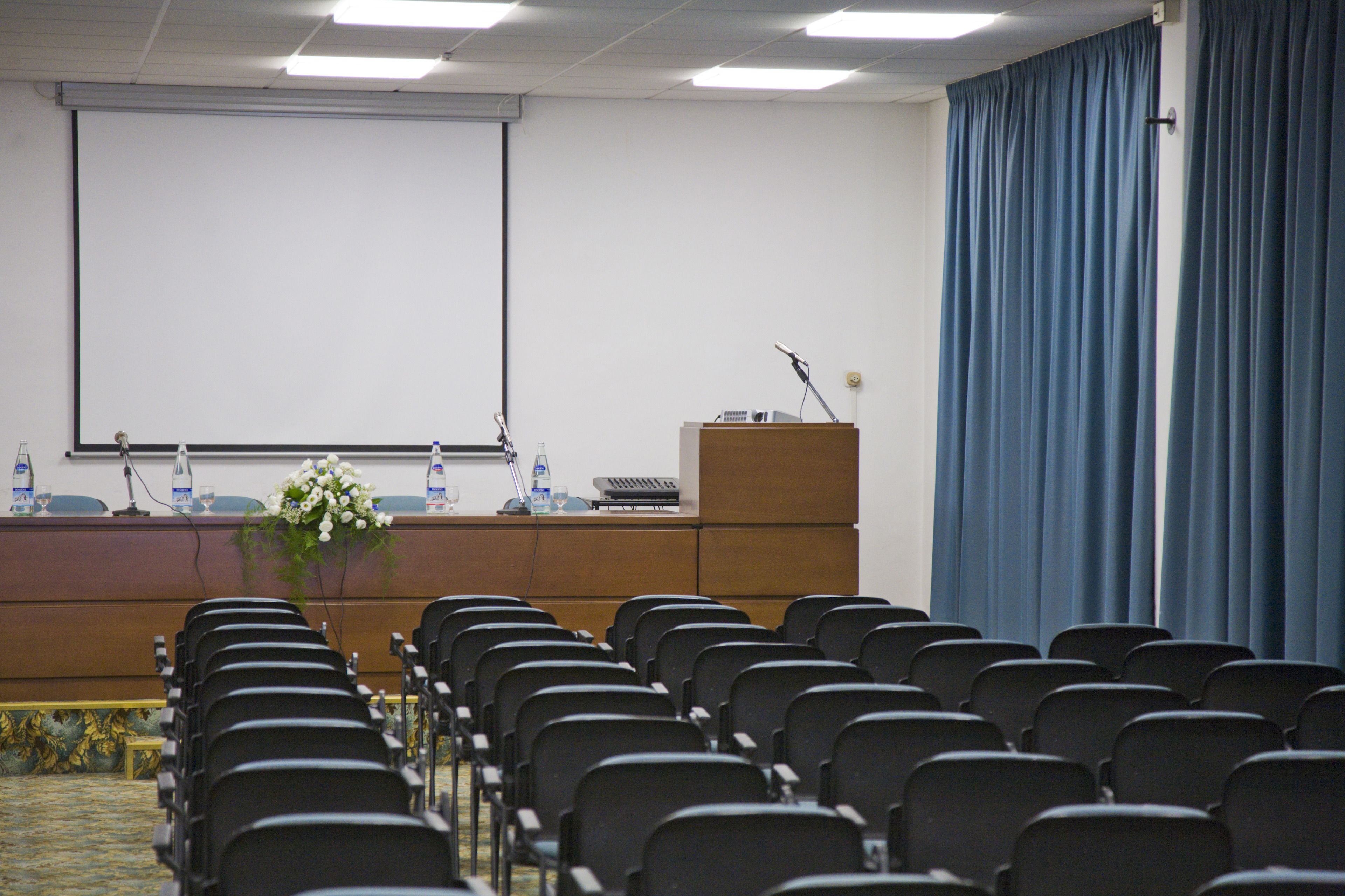 Un salón de conferencias con filas de sillas, un atril, una pantalla de proyector y un dispensador de agua.