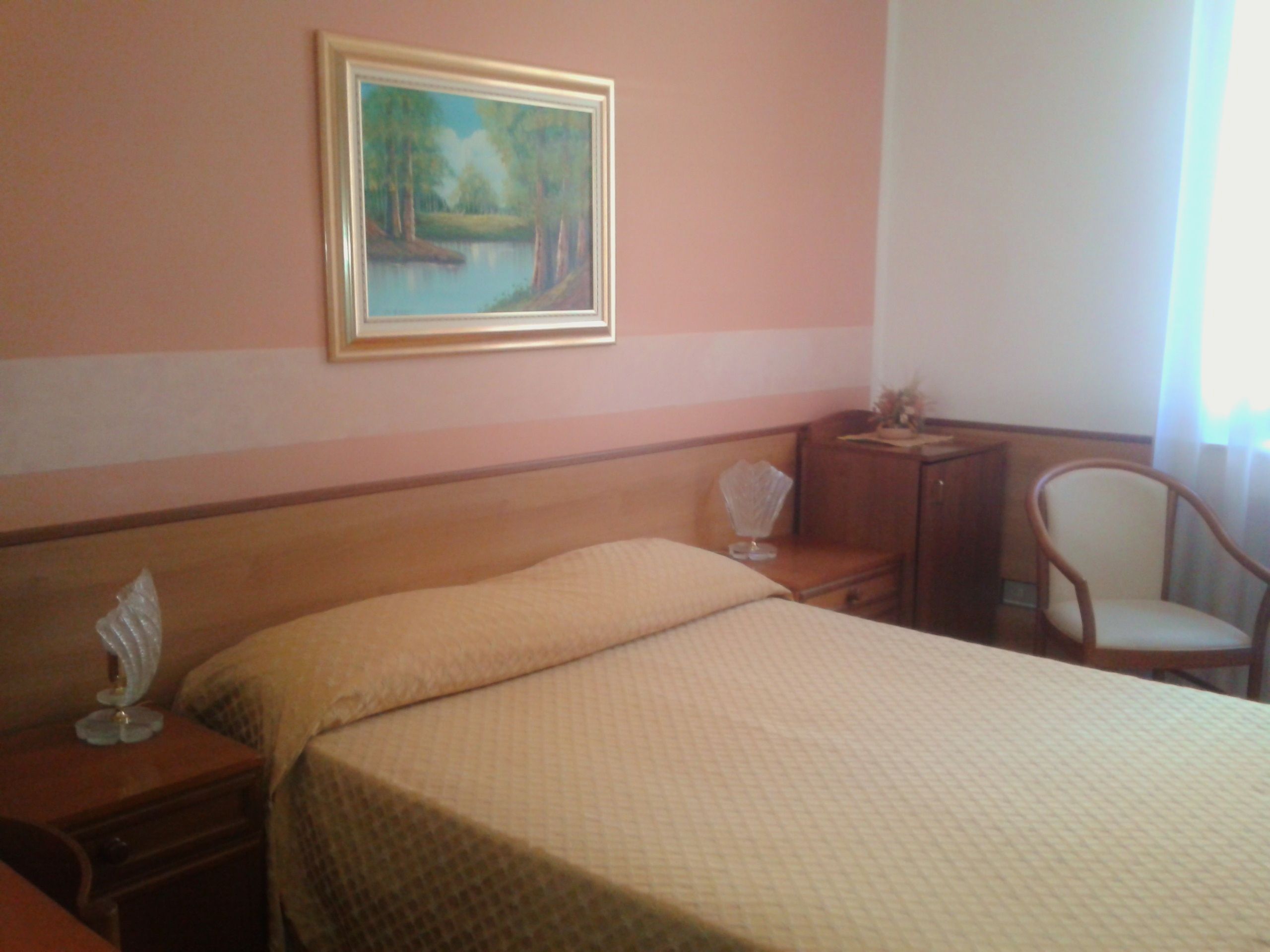 A hotel room with a bed, a nightstand, a chair, a desk, and a painting on the wall.