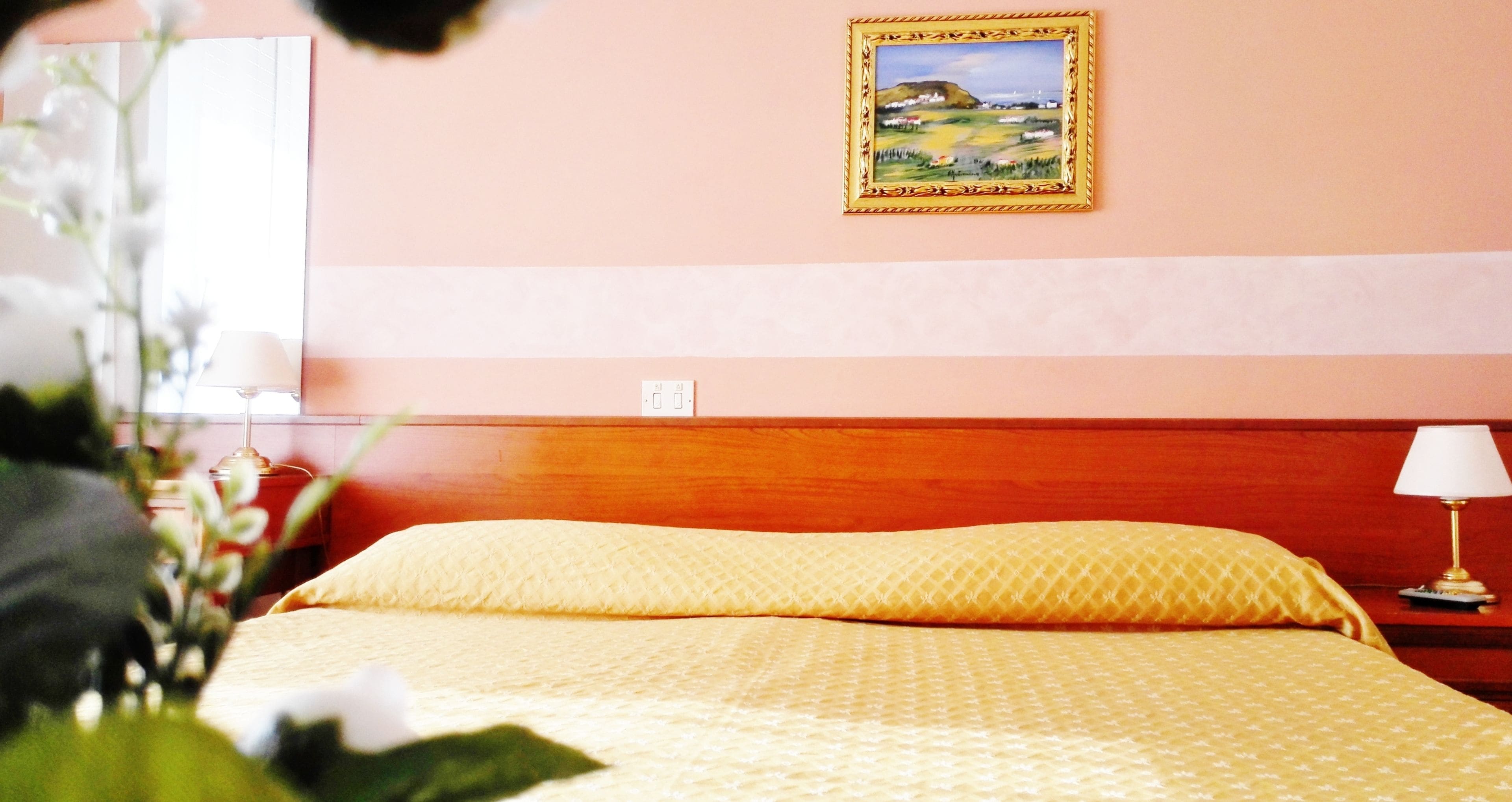 Ein Hotelzimmer mit einem hölzernen Kopfende, einem Bett mit gelbem, gemustertem Bettbezug, einem Gemälde an der Wand und einem Nachttisch mit einer Lampe.