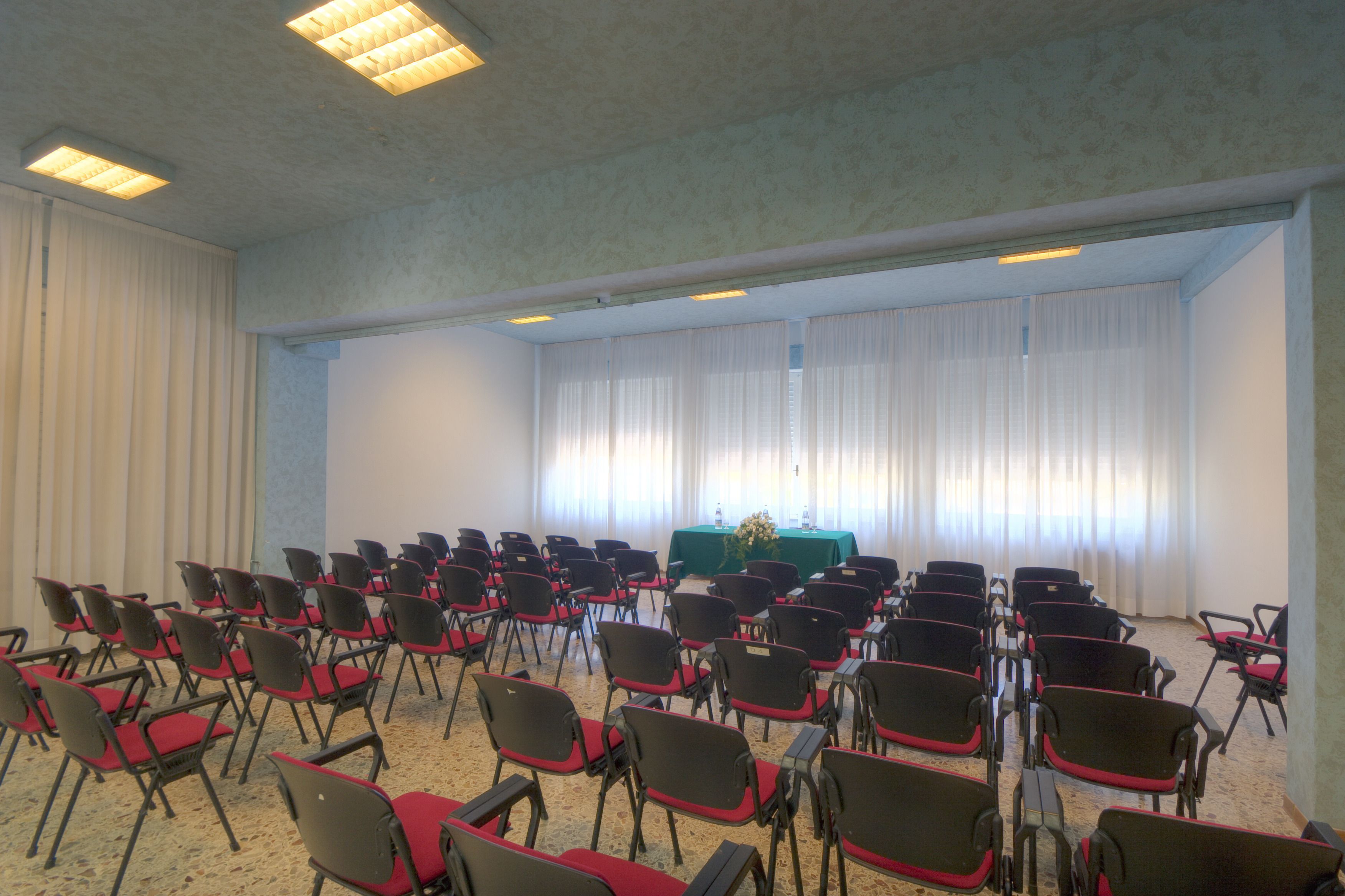 A conference room with rows of chairs, a table with a green tablecloth, and large windows with curtains.