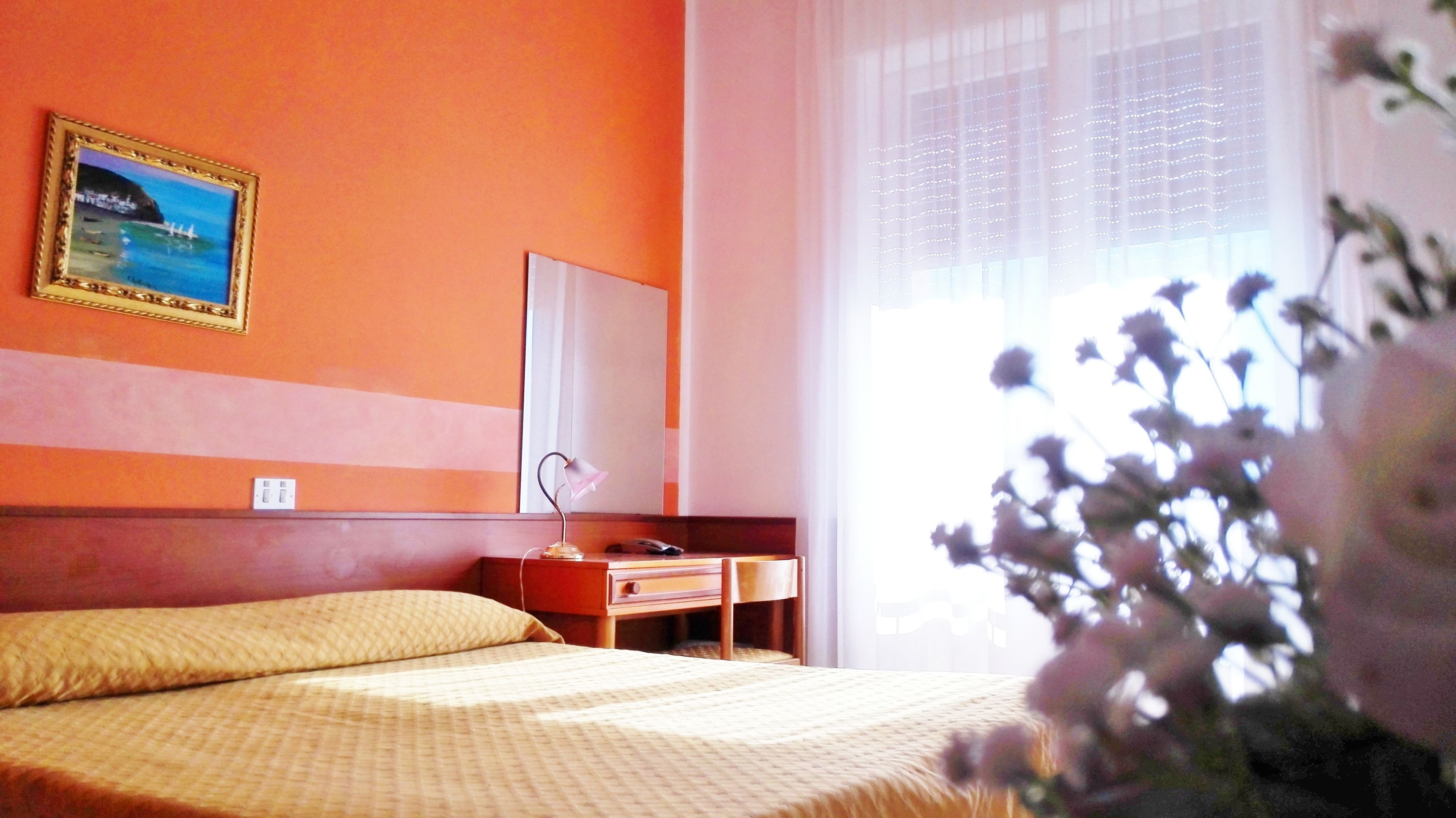 Hotellihuone, jossa on sänky, yöpöytä, peili ja ikkuna, jossa on läpikuultavat verhot.