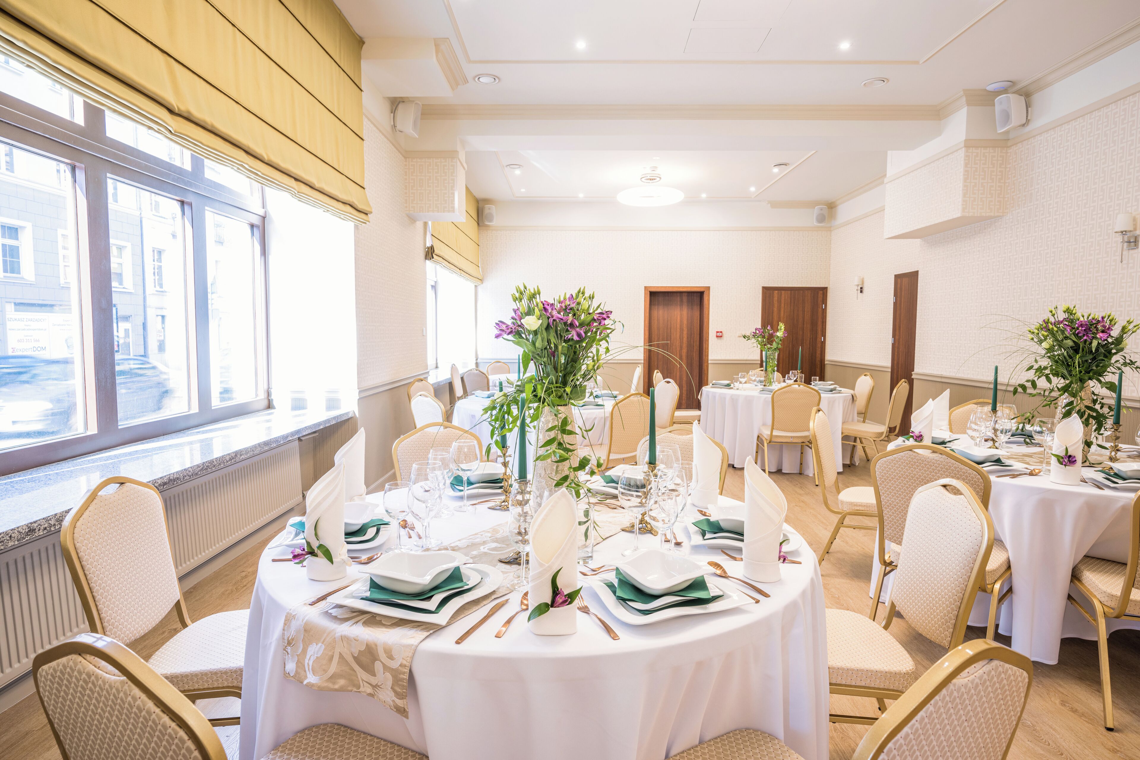 Un salón de banquetes bien iluminado con mesas redondas dispuestas para comer, con manteles blancos, sillas doradas y centros de mesa florales.