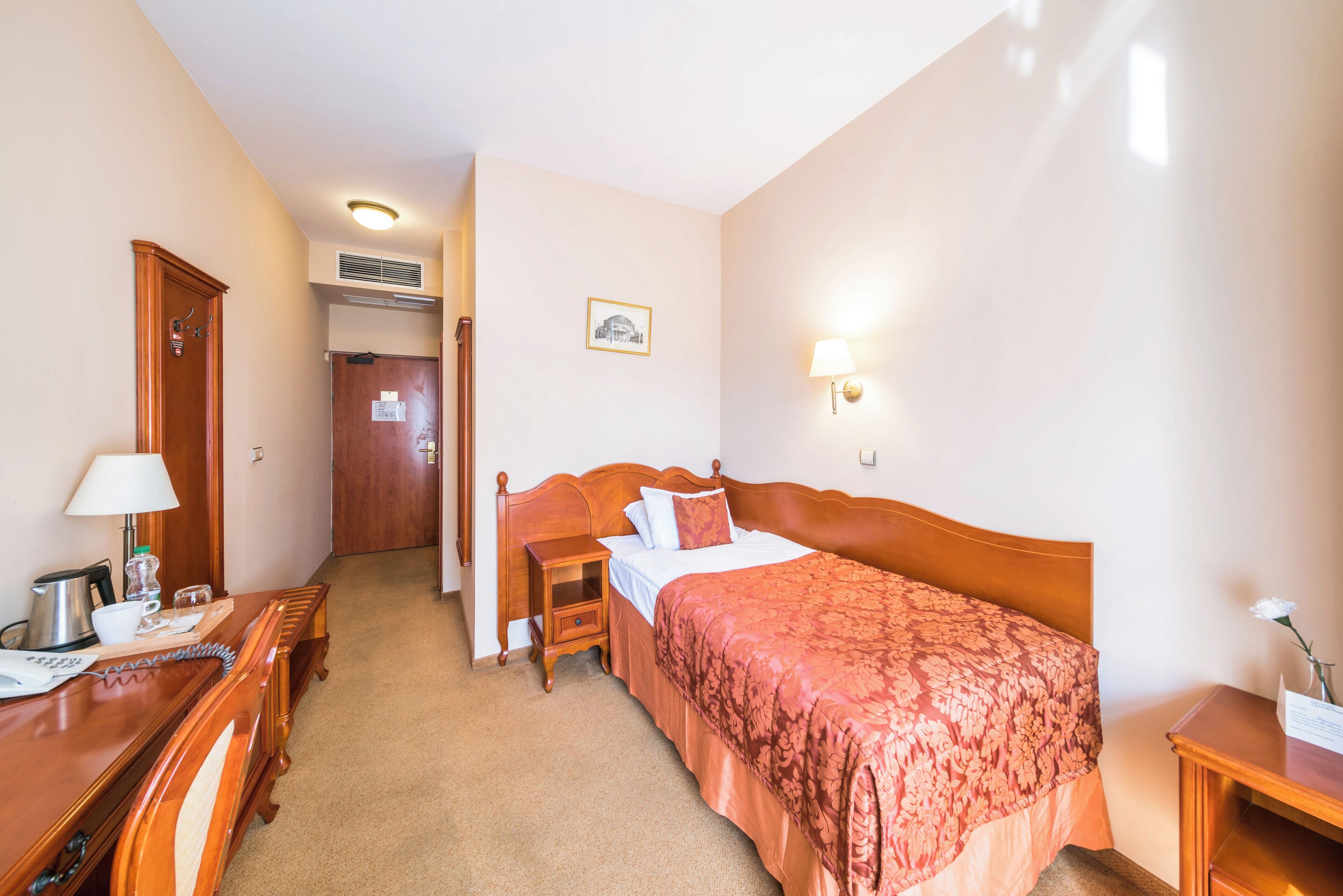 Une chambre d’hôtel avec un lit en bois, un bureau avec une lampe, une table de chevet et une lampe fixée au mur.