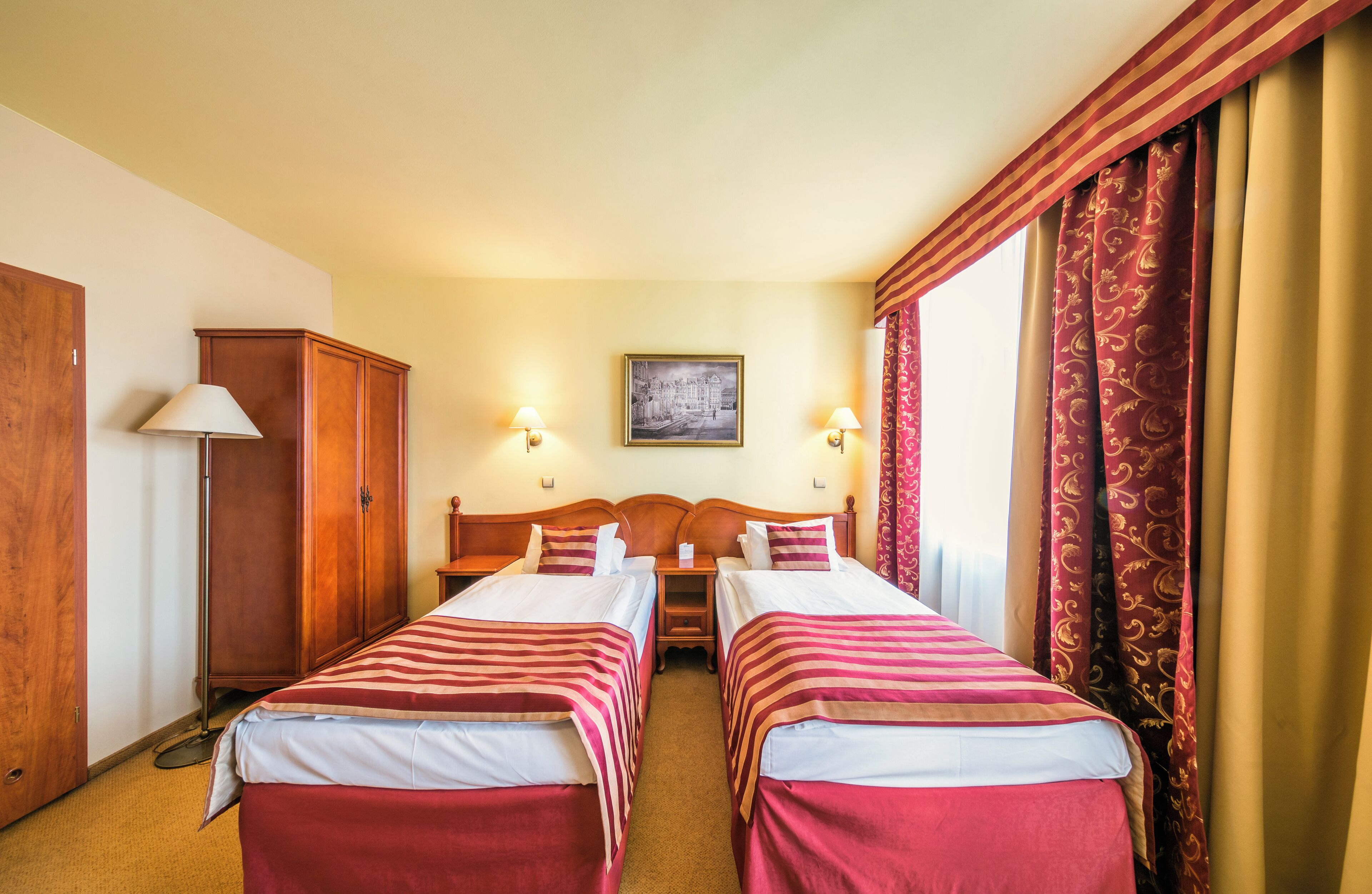 A hotel room with two single beds, a wooden wardrobe, a nightstand, a lamp, and a framed picture on the wall.