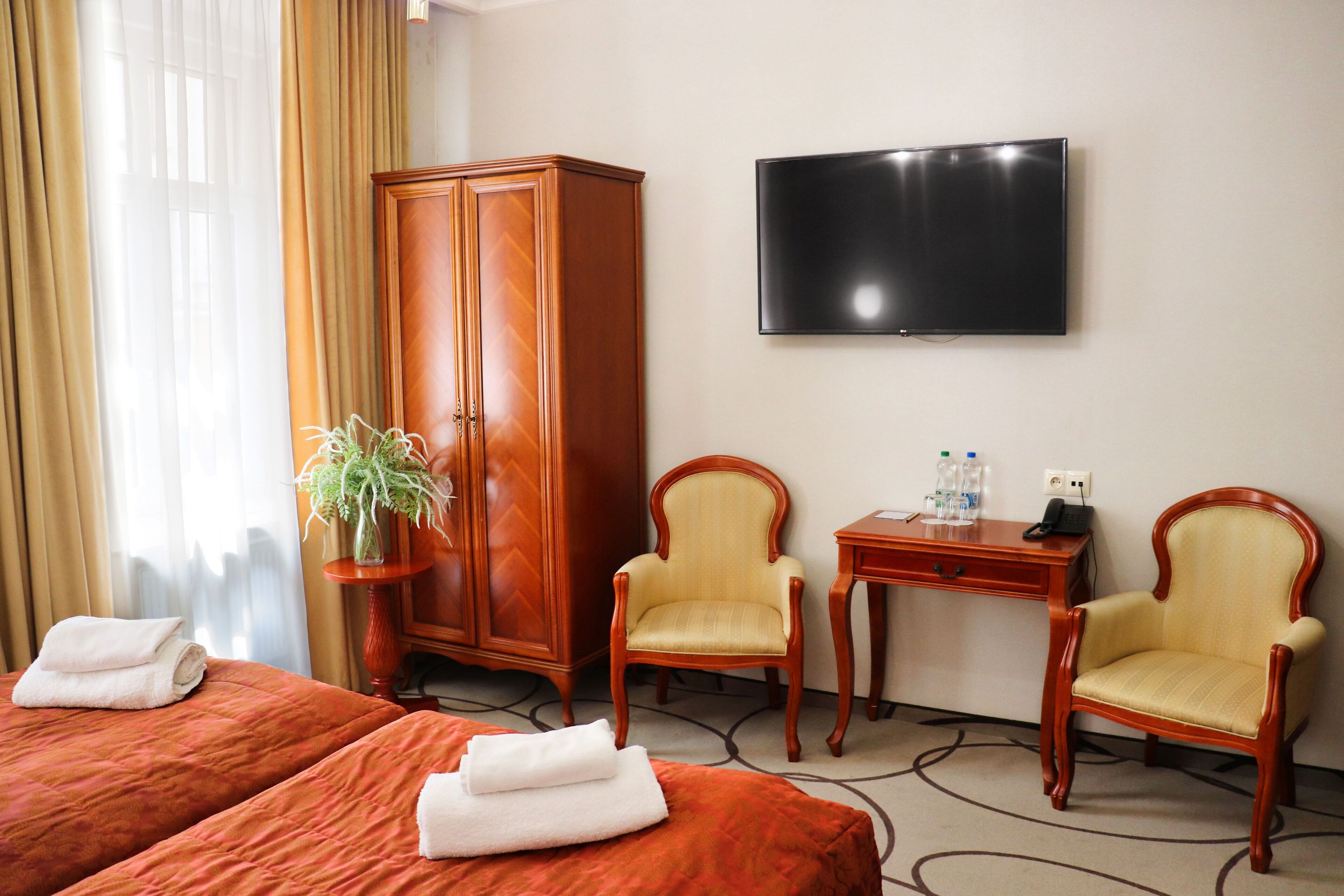 A hotel room with two beds, a TV, a desk, two chairs, a wardrobe, and a potted plant.