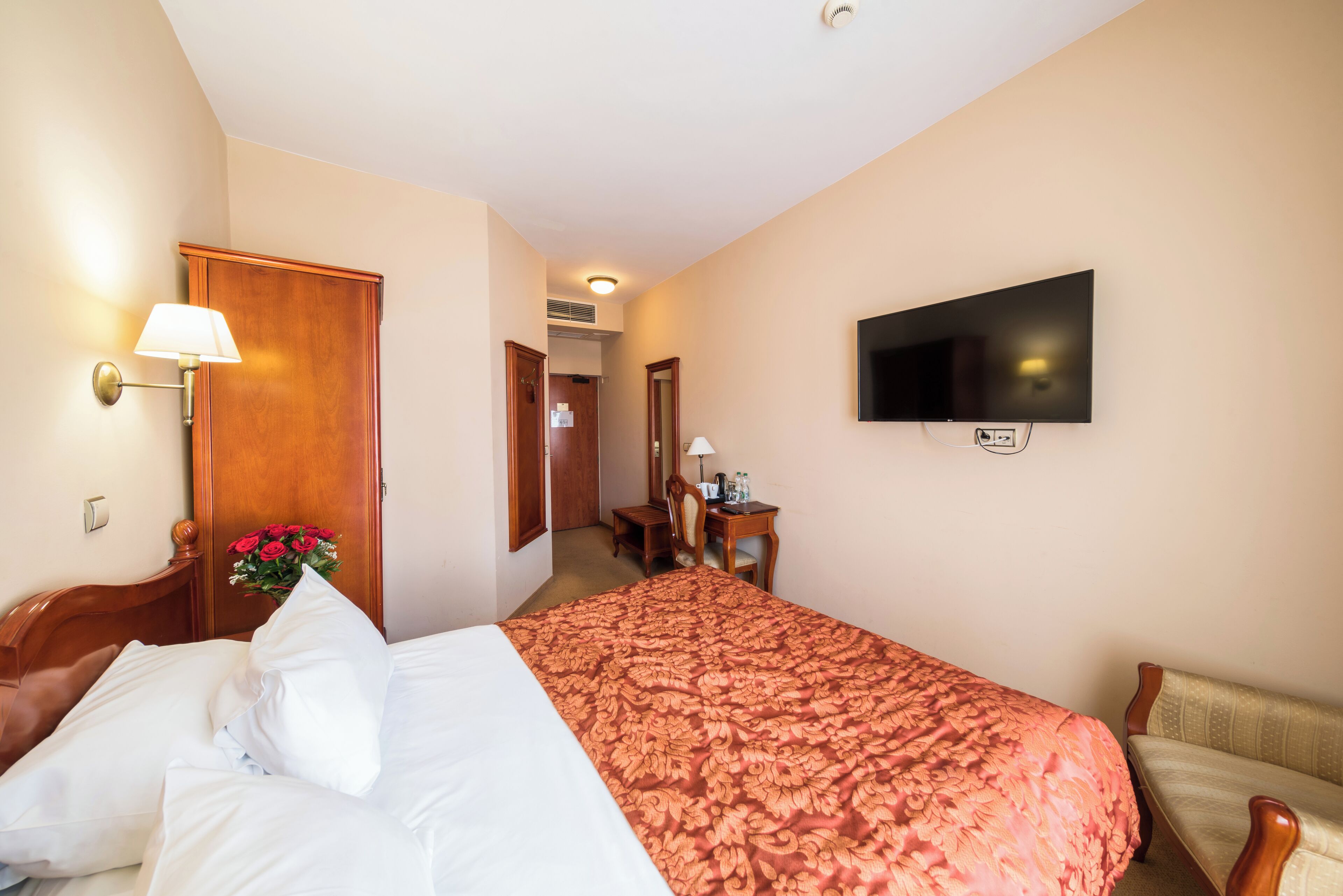 Une chambre d’hôtel comprenant un lit, un bureau avec une chaise, un téléviseur fixé au mur et une porte en bois.
