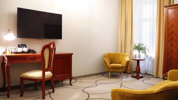 Quarto de hotel com televisão de tela plana na parede, uma escrivaninha de madeira com abajur, uma poltrona amarela, um sofá e uma mesa pequena com uma planta.