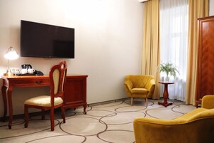 Quarto de hotel com televisão de tela plana na parede, uma escrivaninha de madeira com abajur, uma poltrona amarela, um sofá e uma mesa pequena com uma planta.