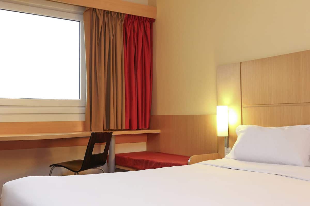 A hotel room with a bed, a desk, a chair, a window with curtains, and a bedside lamp.