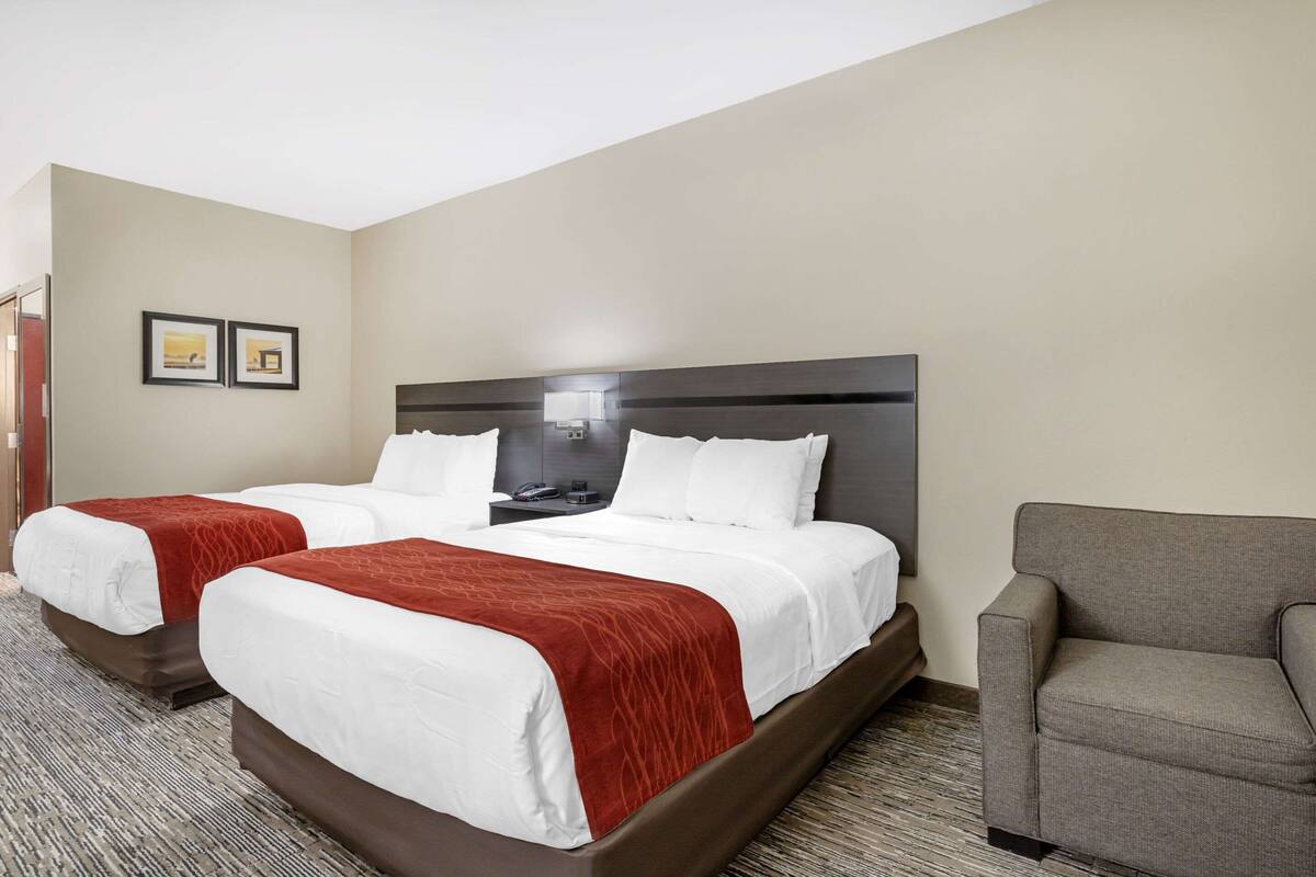 A hotel room with two beds, a gray armchair, and a nightstand with a lamp.
