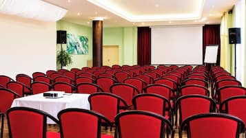 Un salón de conferencias con filas de sillas rojas, una mesa blanca y un proyector, y una pantalla grande.
