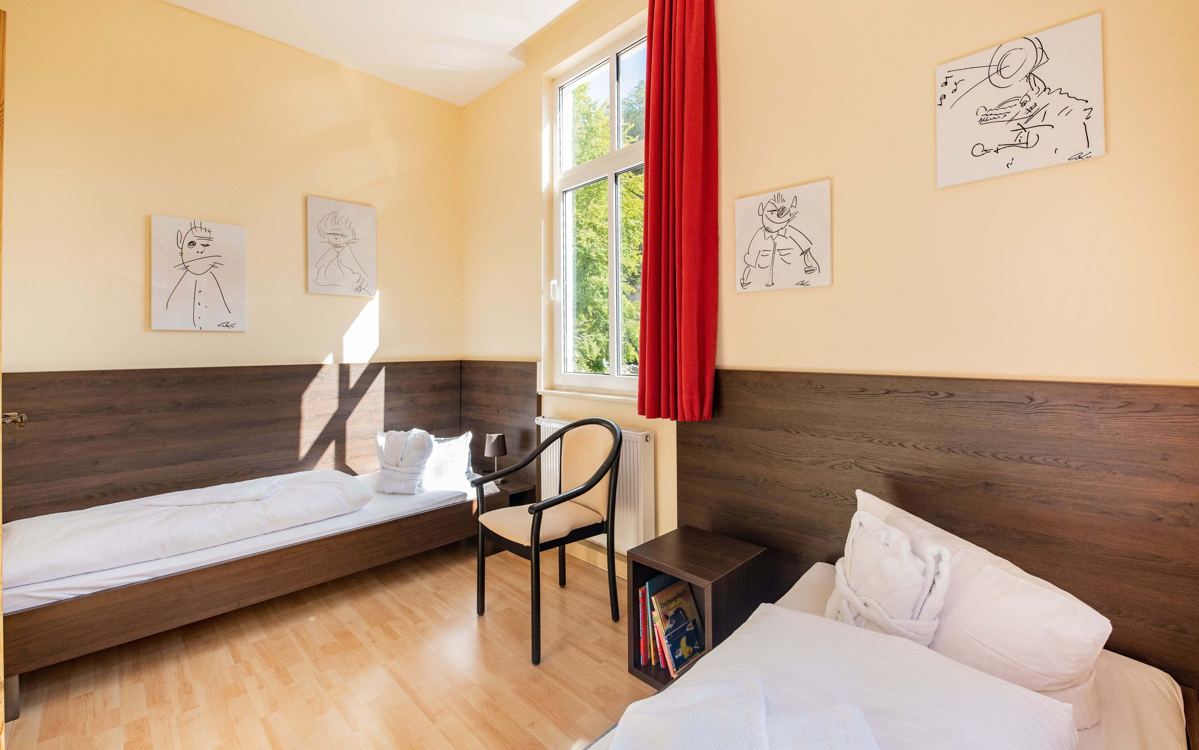 Une chambre d’hôtel avec deux lits simples, un plancher en bois, une fenêtre avec des rideaux rouges et des œuvres d’art murales.