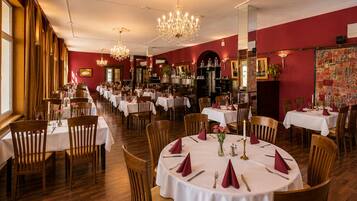 A restaurant with round tables, wooden chairs, chandeliers, and red walls.