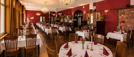 A restaurant with round tables, wooden chairs, chandeliers, and red walls.