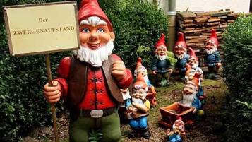 Un jardín con estatuas de gnomos, incluyendo uno que sostiene un cartel que dice "Der ZWERGENAUFSTAND".