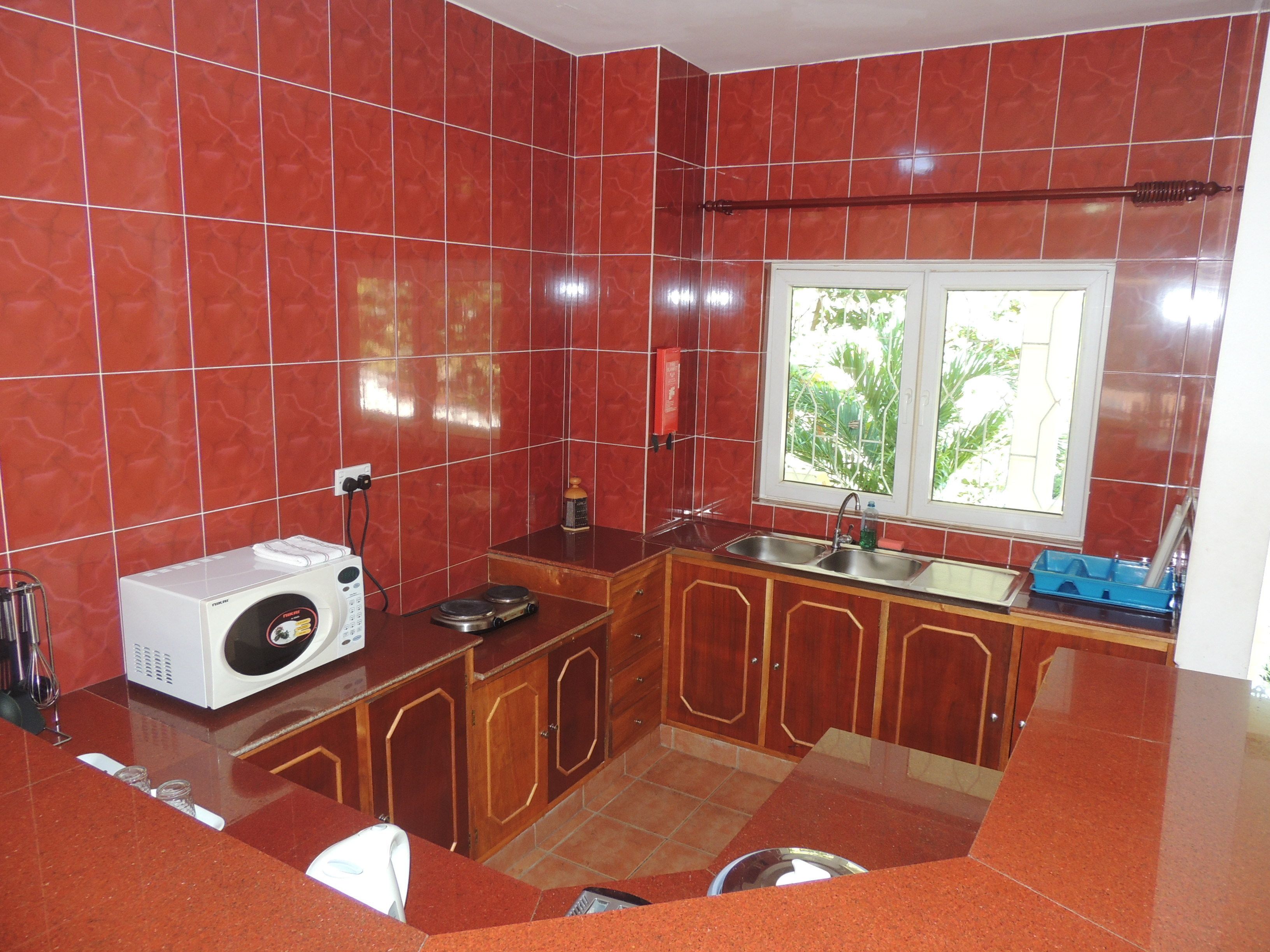 A kitchen with red tiles, wooden cabinets, a built-in microwave, a sink, and a window with a view of greenery.