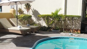 Una piscina con sillas de descanso y sombrillas, rodeada de plantas en macetas y una pared.