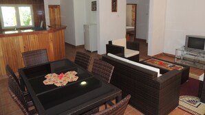Un comedor con mesa negra y sillas de mimbre, una cocina con refrigerador, y una sala de estar con televisor y sofá.
