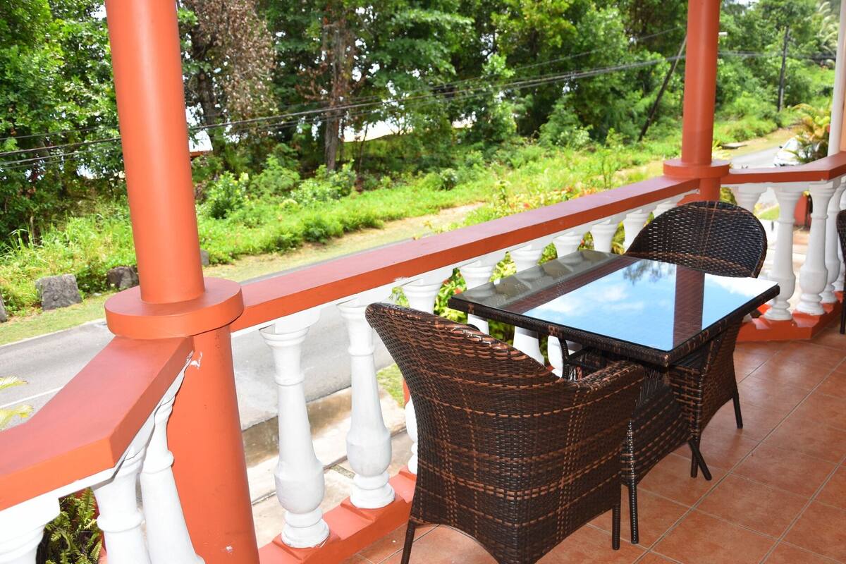 A balcony with wicker chairs and a glass table, overlooking a lush garden and a road.