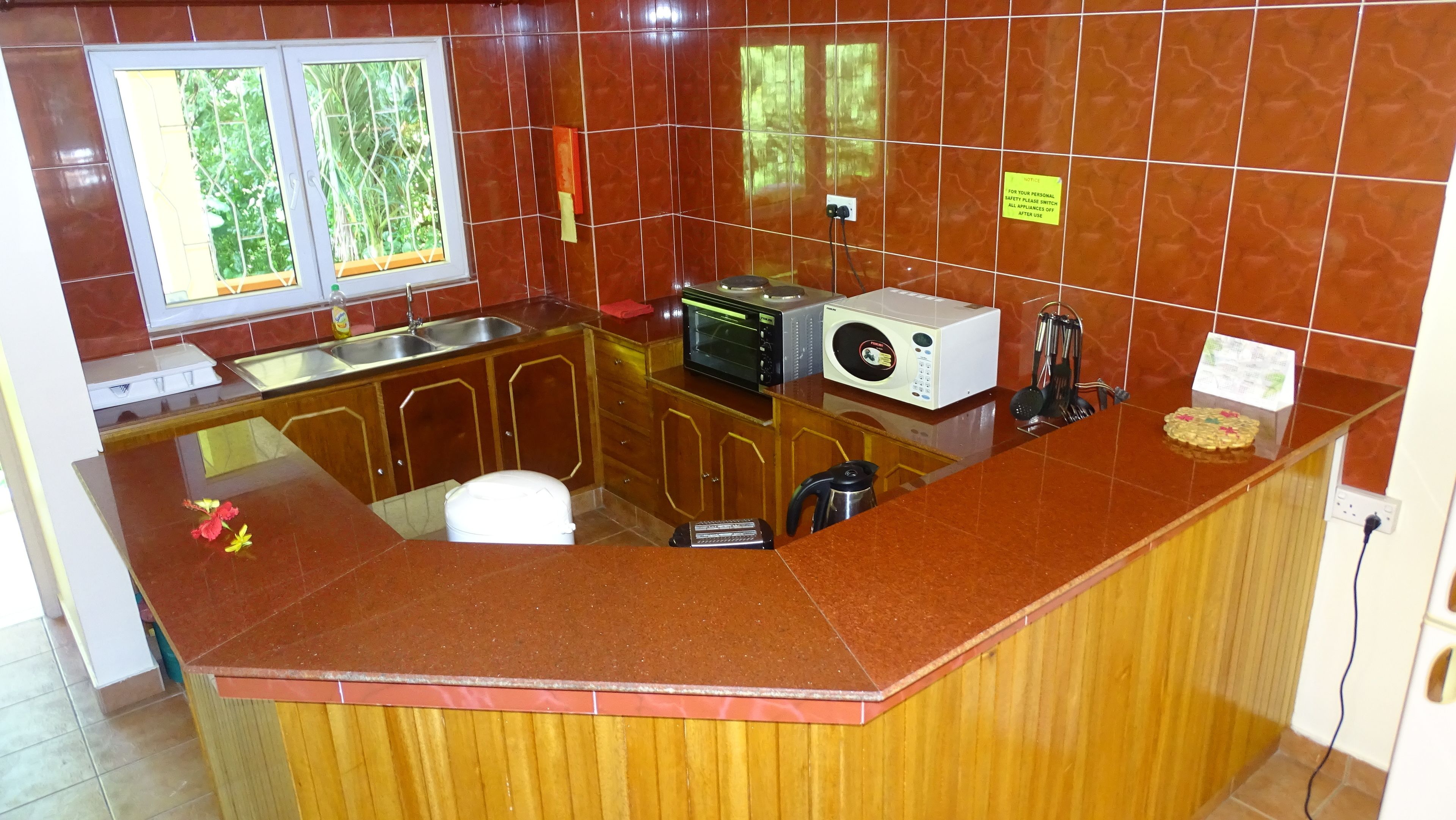 A kitchen with a red countertop, wooden cabinets, a sink, a microwave, a toaster, and a window with a view of greenery.