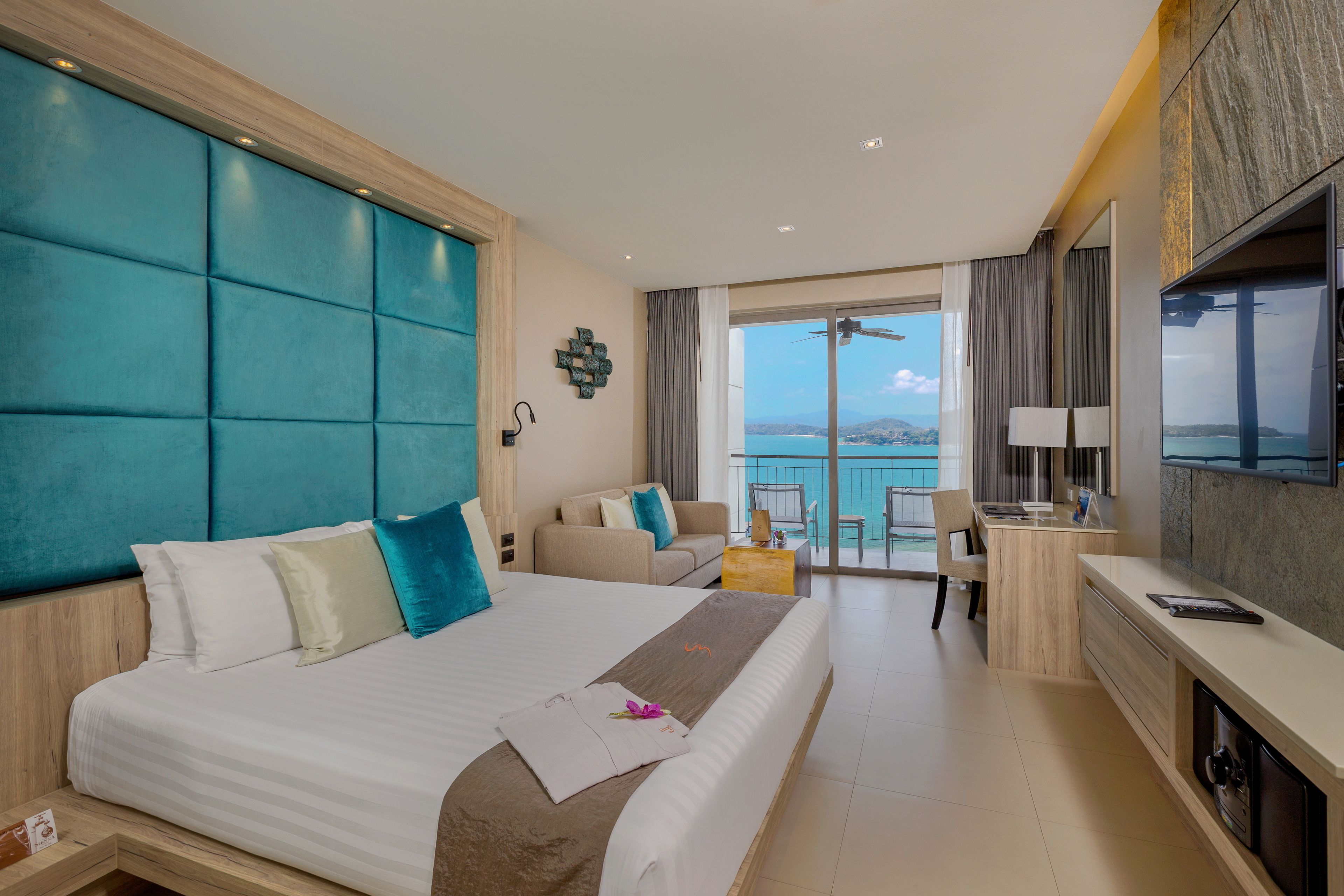 A modern hotel room with a large bed, a sofa, a desk, and a balcony with a view of the sea.