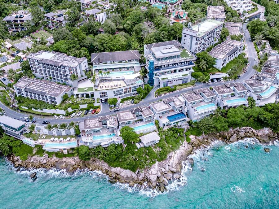 An aerial view of a coastal resort with multiple buildings, including a hotel with a pool, surrounded by lush greenery and the ocean.
