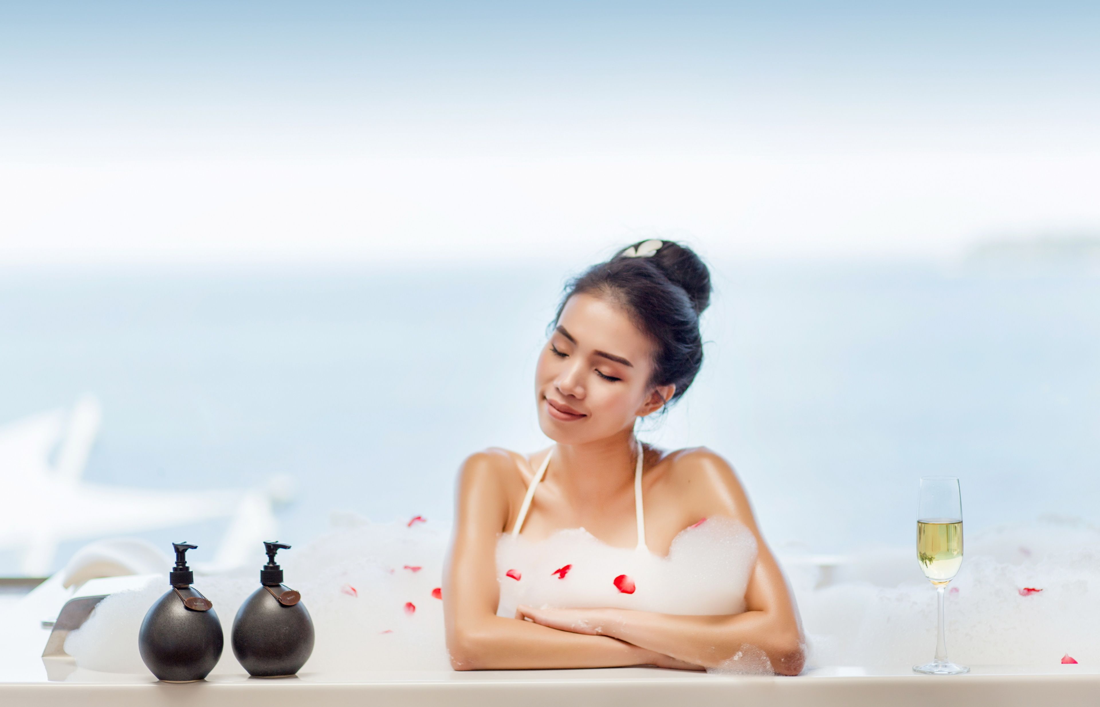 A person in a bathtub with rose petals, a bottle of lotion, and a glass of wine.