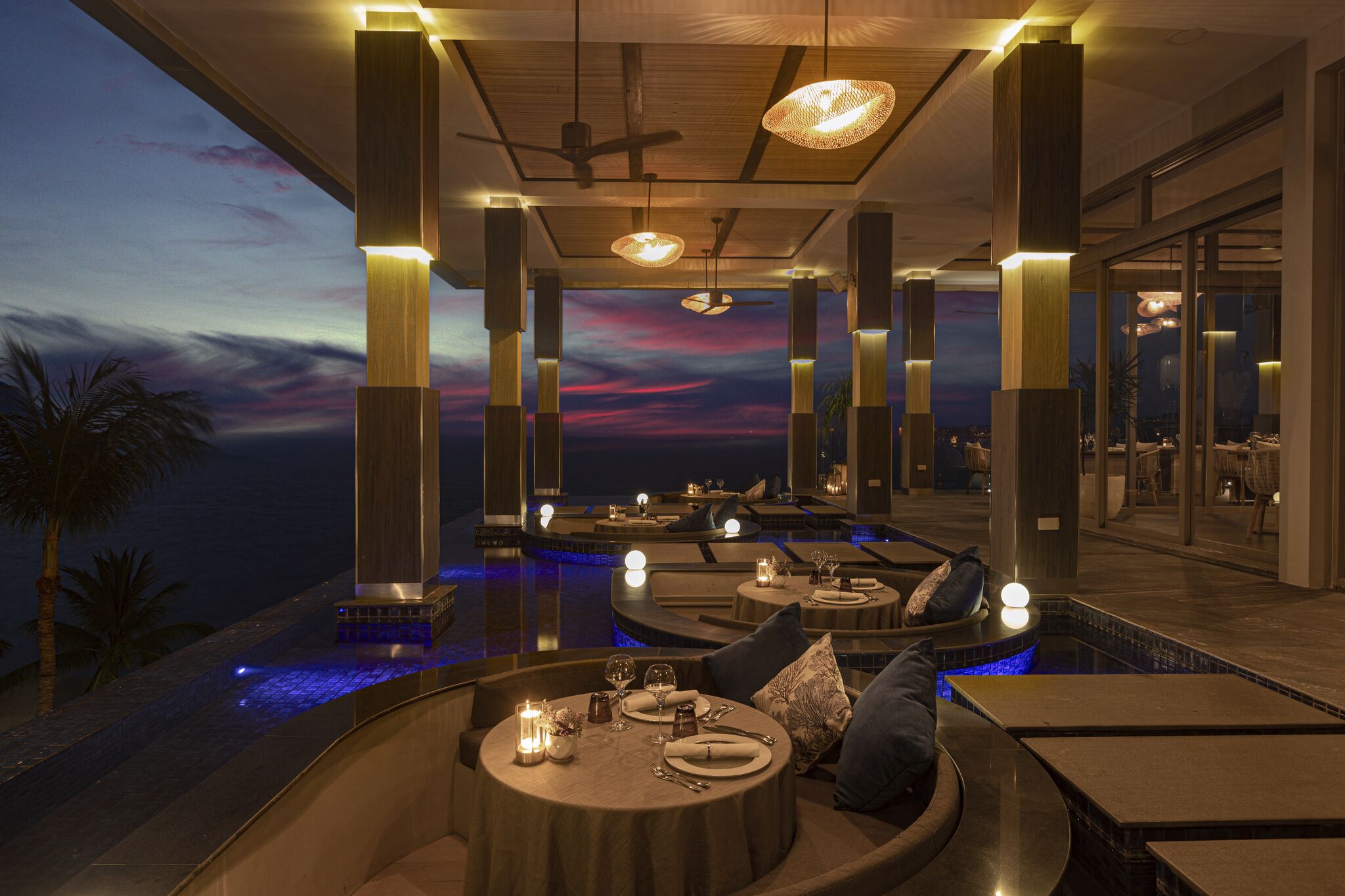Un espace extérieur luxueux aménagé pour les repas, avec des tables rondes, des bougies et une vue imprenable sur le coucher du soleil.