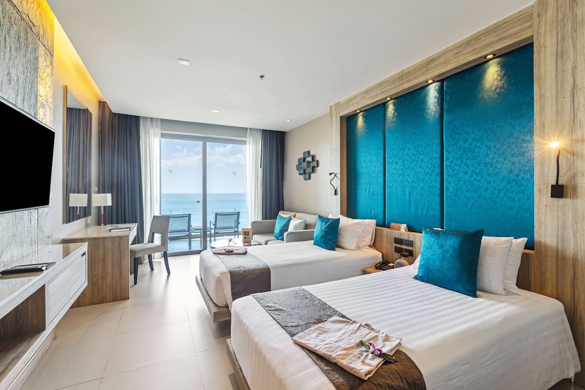 A hotel room with two beds, a desk, a TV, and a view of the sea.