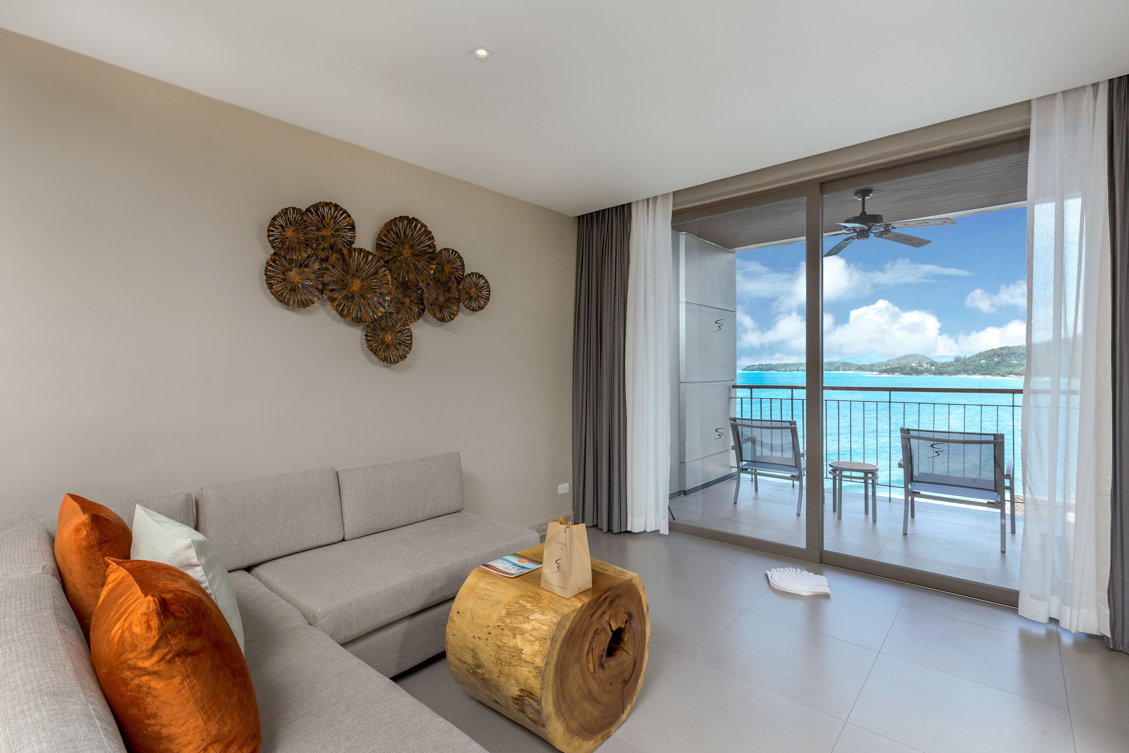 A modern living room with a grey sofa, decorative wall art, and a large wooden coffee table. A balcony with a view of the sea is visible through the glass doors.