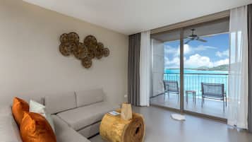 A modern living room with a grey sofa, decorative wall art, and a large wooden coffee table. A balcony with a view of the sea is visible through the glass doors.