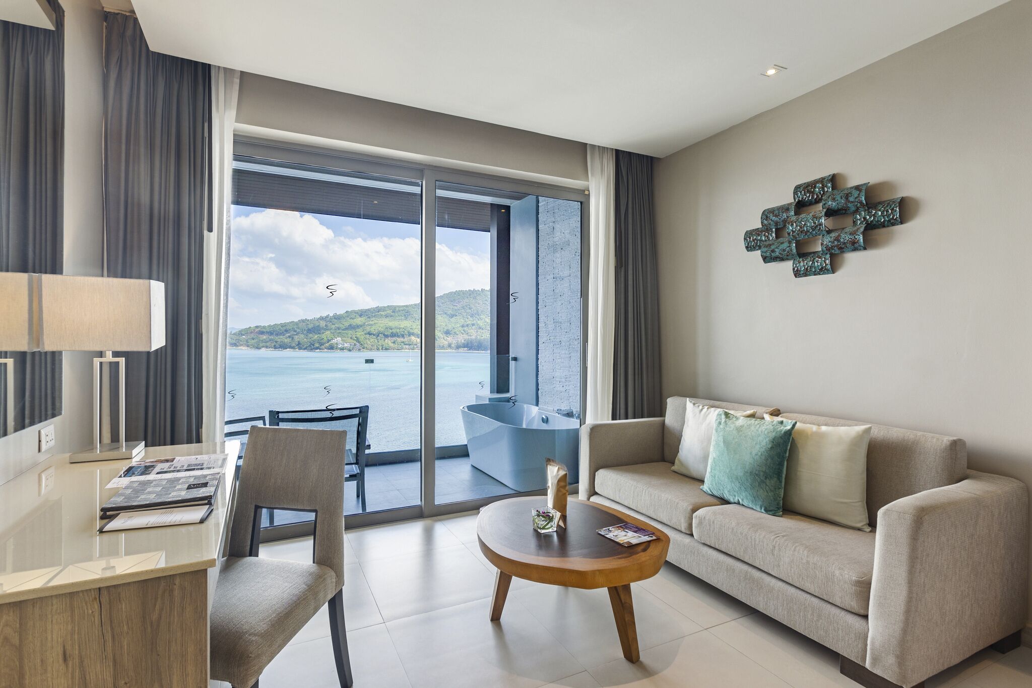 A modern hotel room with a large sliding glass door leading to a balcony with a view of the sea and hills. There is a sofa, a small round table, and a desk with a lamp.