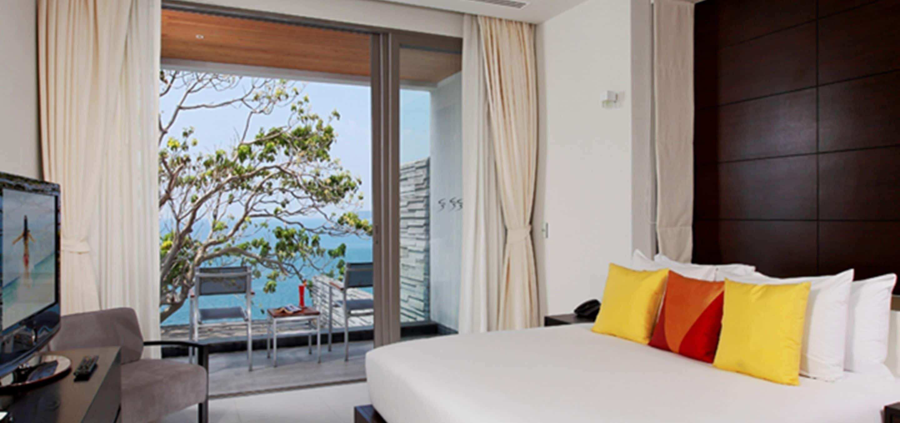 A hotel room with a large bed, a desk with a computer, and a view of the sea through a sliding glass door.