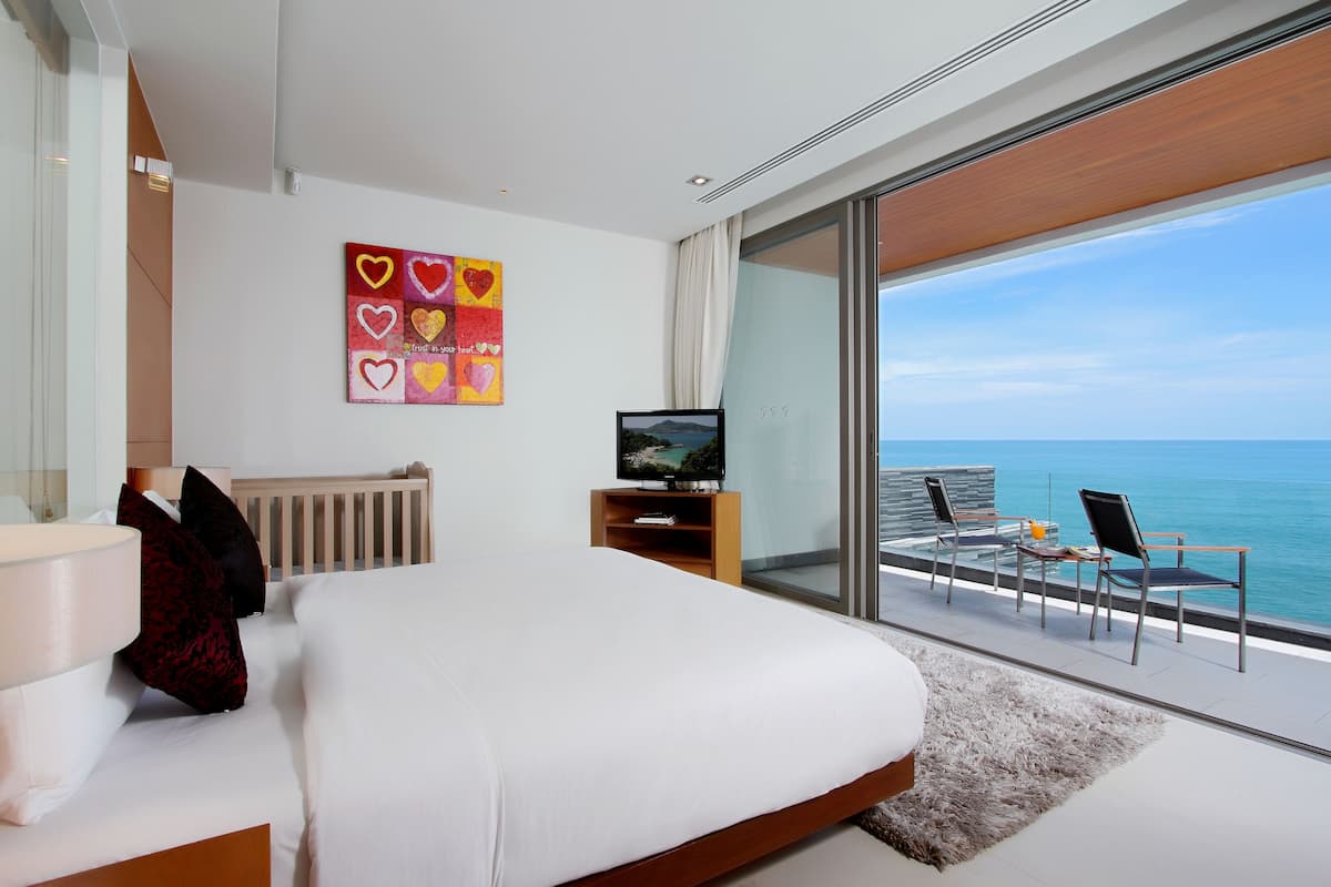 Habitación de hotel moderna con una cama grande, televisión, vista al océano y balcón con sillas y una mesa.