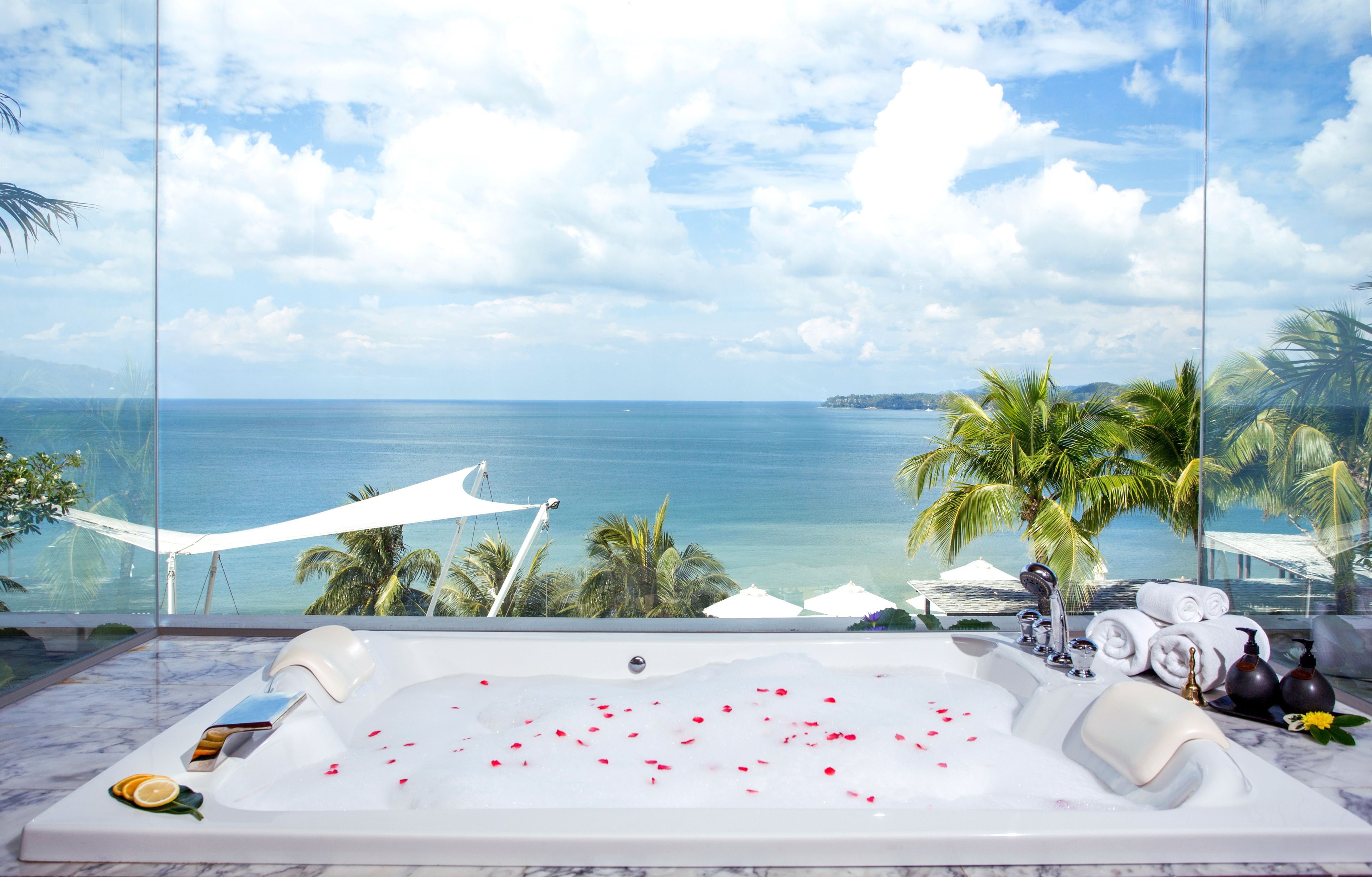 Una vasca da bagno lussuosa con petali di rosa, asciugamani e articoli da toilette, con vista su una spiaggia con palme e l'oceano.