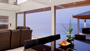 Un salon moderne doté d’une grande baie vitrée donnant sur la mer. On y trouve un canapé, une table à manger avec un panier de fruits, ainsi qu’un petit espace jardin avec une plante.