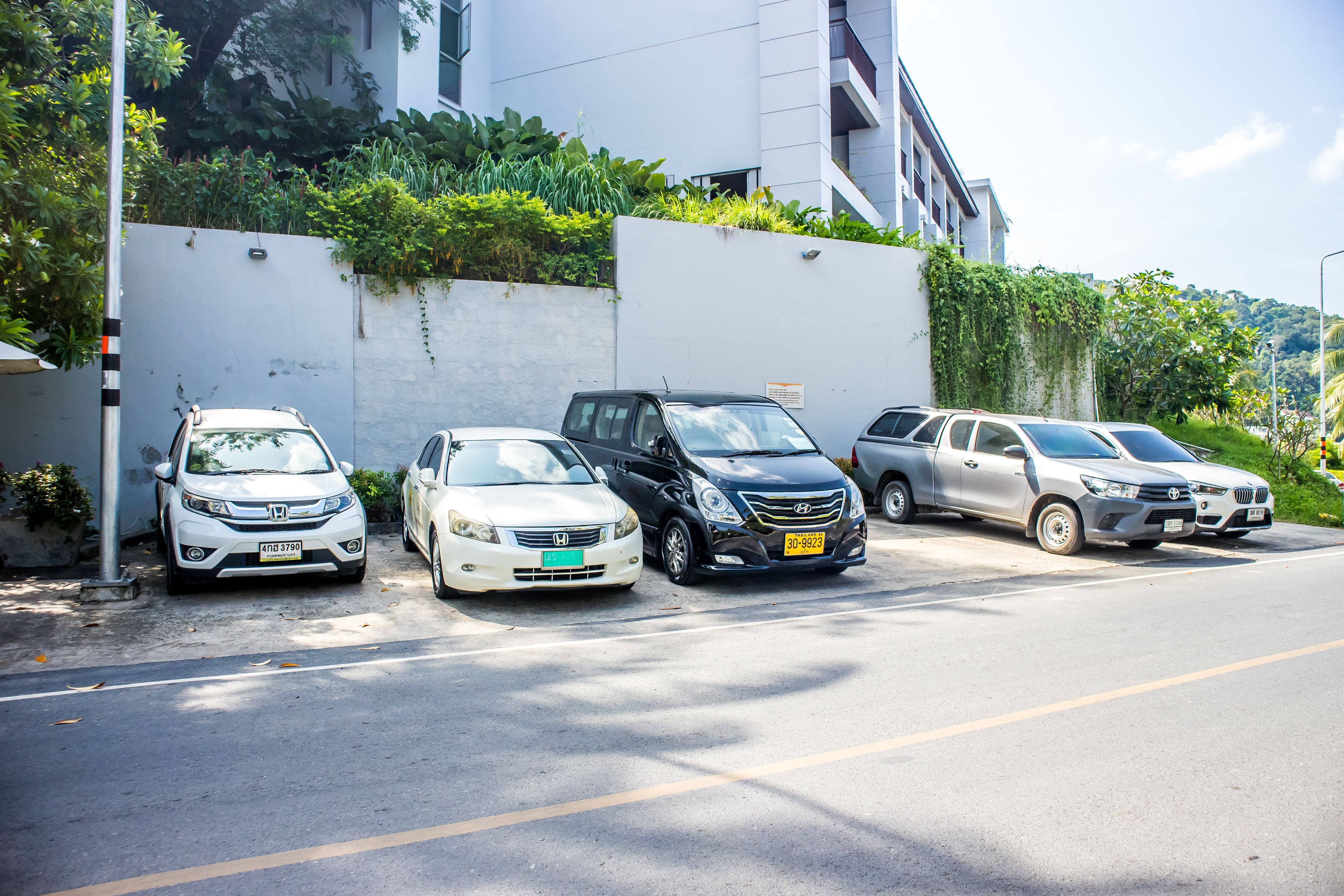 A row of parked cars along a street with a white building and greenery in the background.