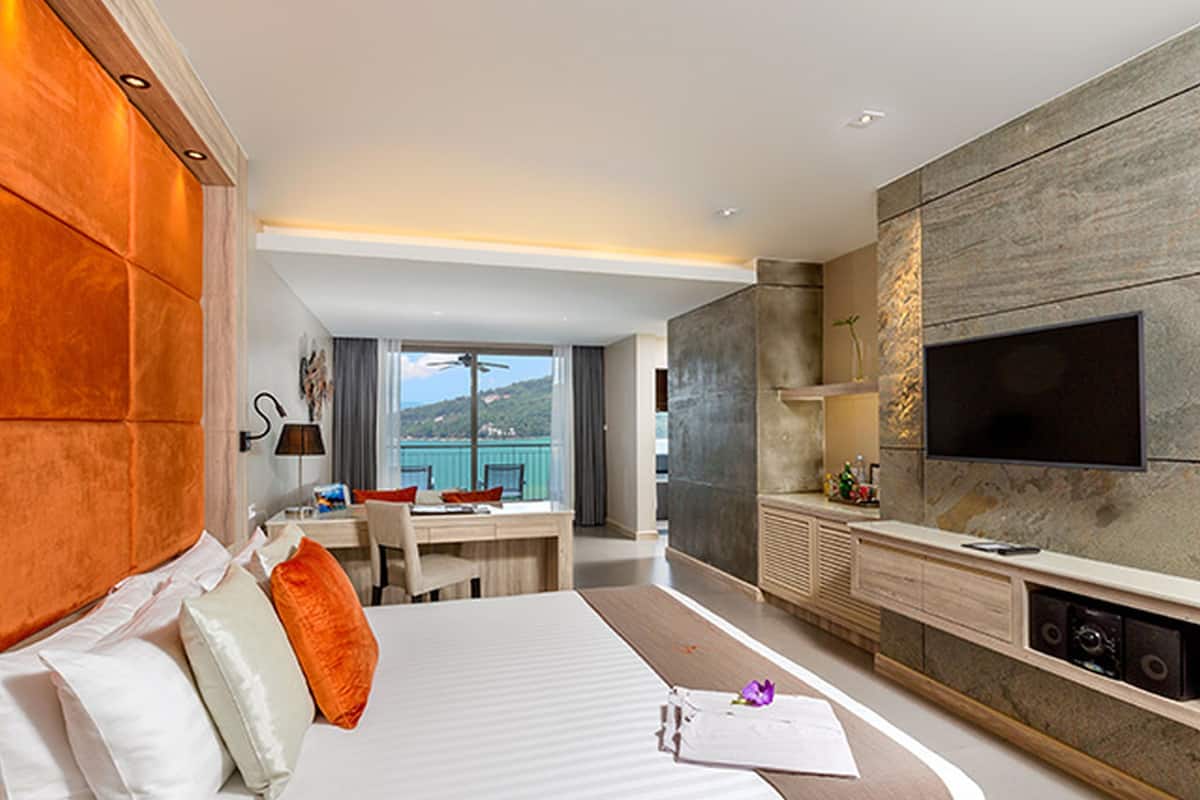A hotel room with a bed, a desk, a TV, and a view of the sea.