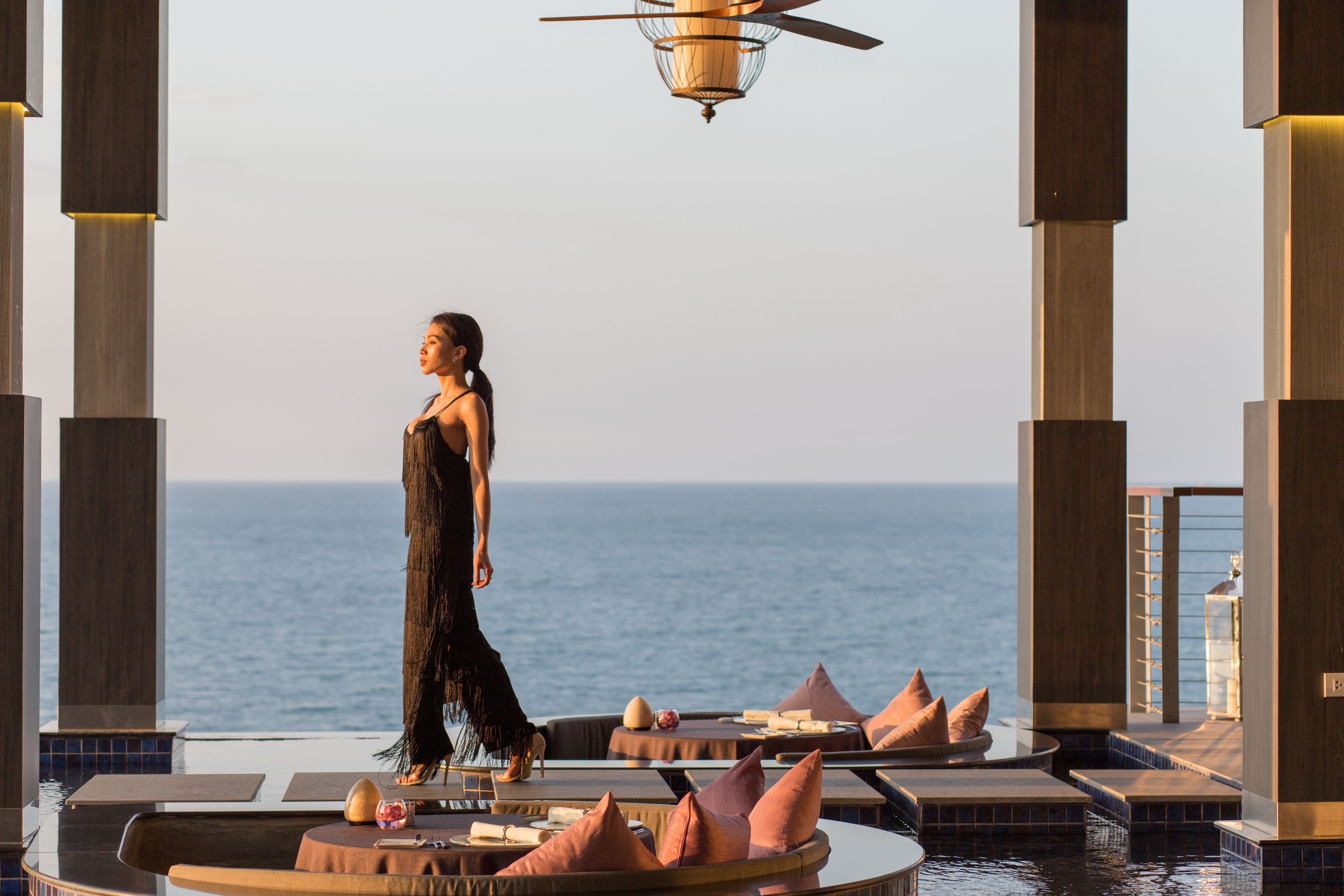 Eine Frau in einem langen Kleid geht über einen Pooldeck mit Blick auf das Meer.