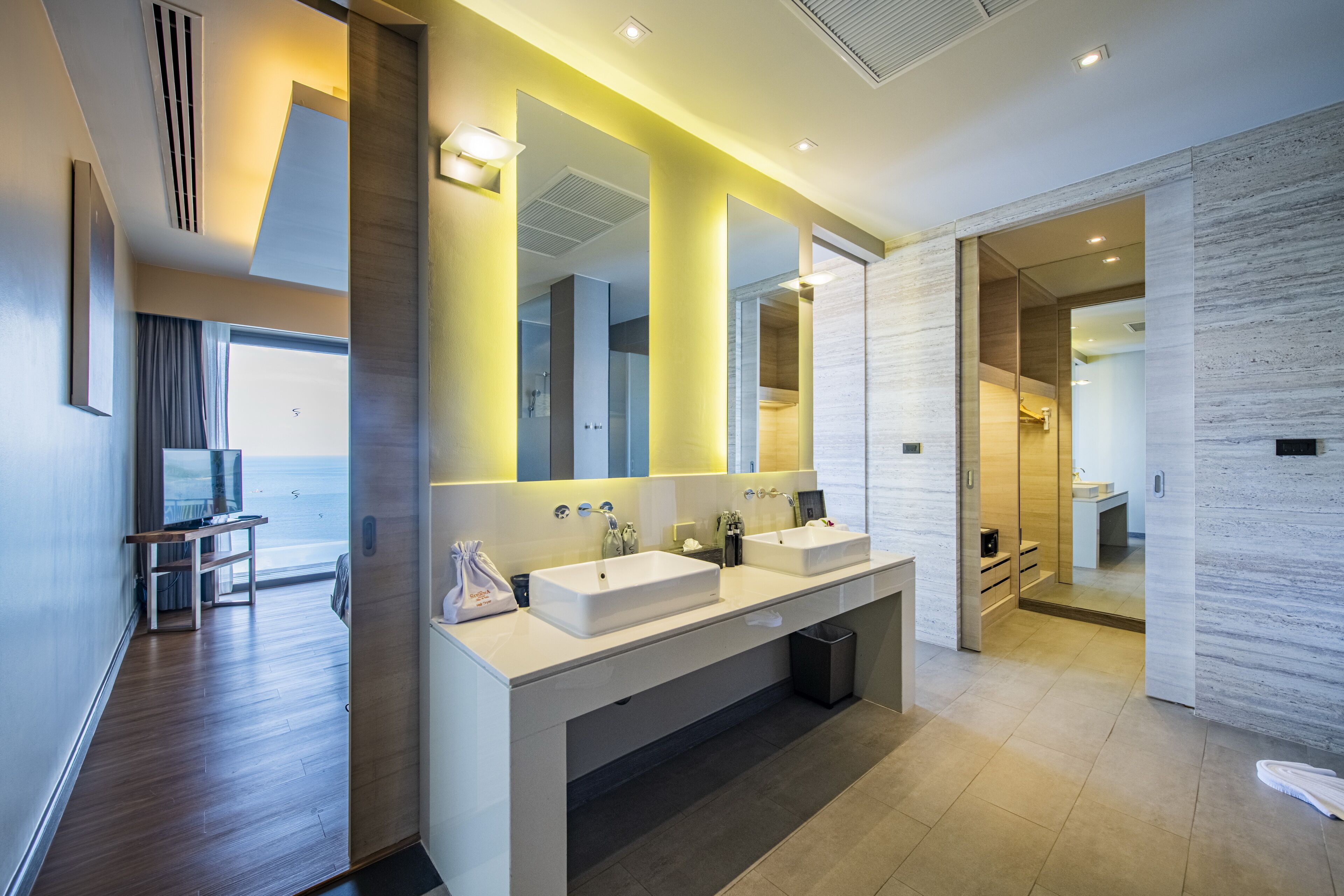 A modern bathroom with dual sinks, a large mirror, and a view of the sea.