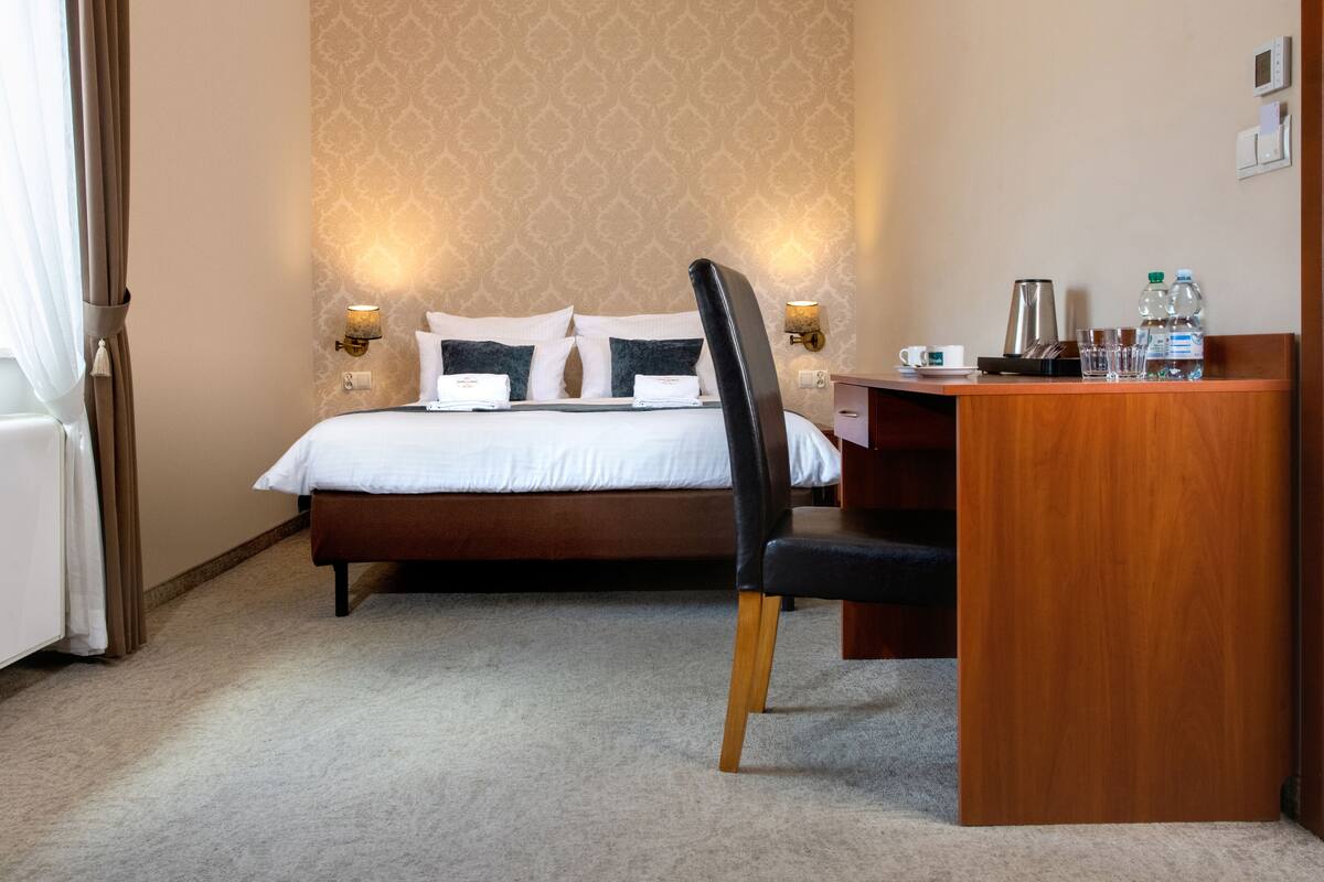 A hotel room with a bed, a desk, a chair, and a nightstand with a kettle, cups, and bottles.