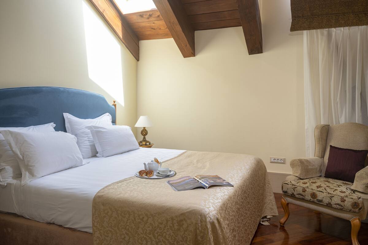 A neatly made bed with white linens, a side table with a lamp, and a chair with a floral pattern.