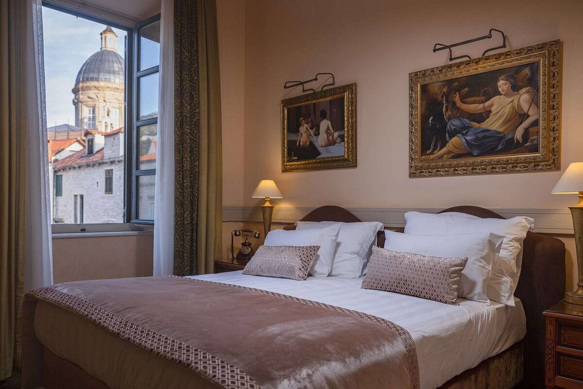 A hotel room with a large bed, two paintings on the wall, a window with a view of a building, and a nightstand with a lamp.