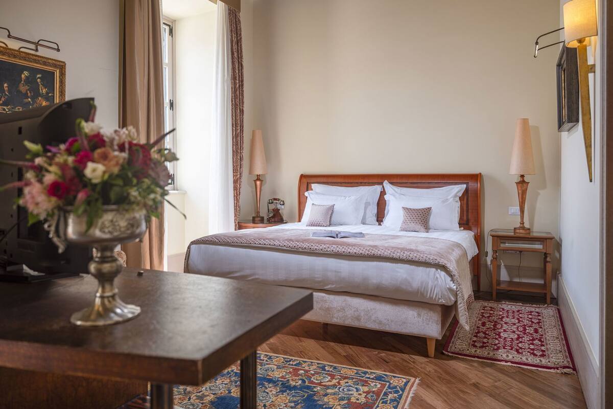 A neatly arranged bedroom with a wooden bed, bedside lamps, a table with a vase of flowers, and a patterned rug.
