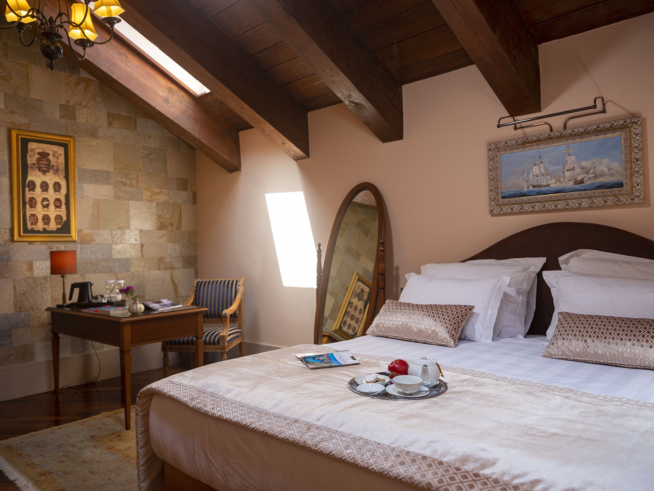 A bedroom with a large bed, a wooden ceiling, a stone wall, a desk with a chair, and a painting on the wall.