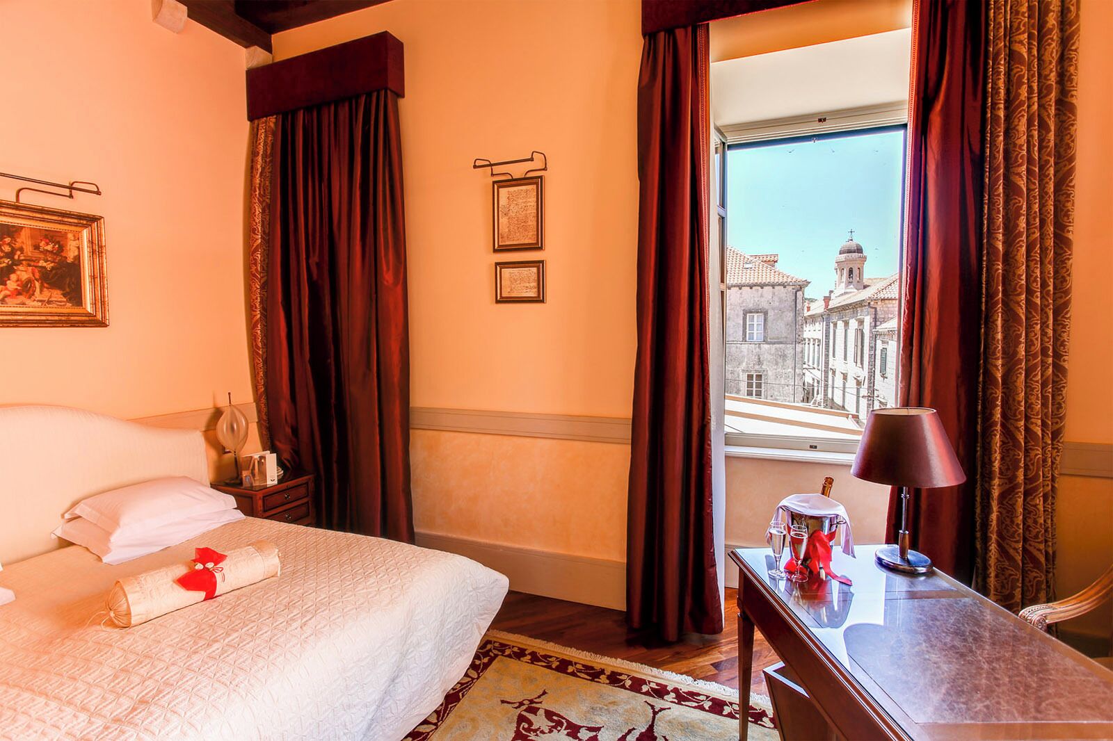 A hotel room with a bed, a desk with a lamp, a window with curtains, and framed pictures on the wall.