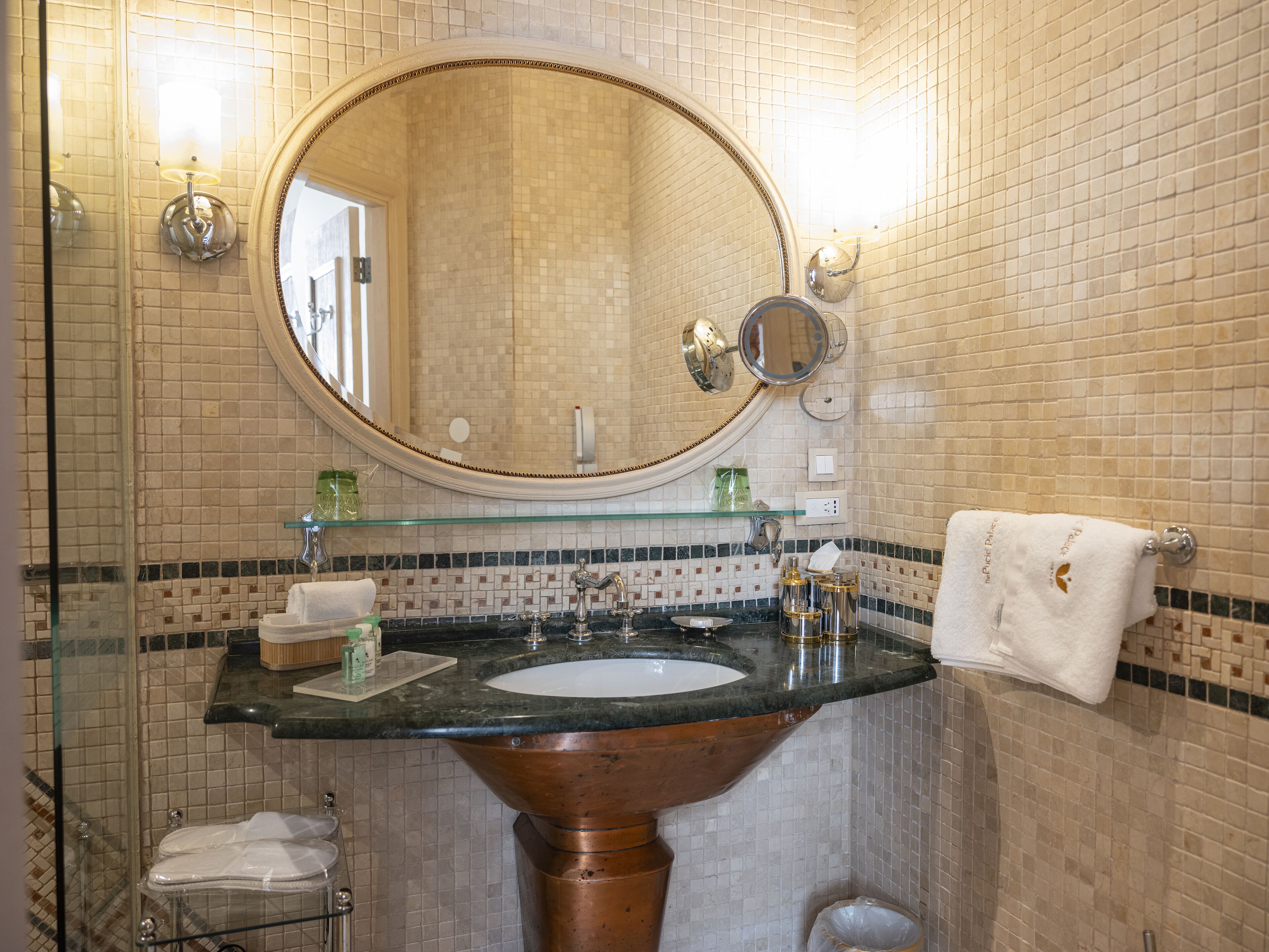 A bathroom with a large oval mirror, a sink with a copper pedestal, and a glass shelf above the sink.
