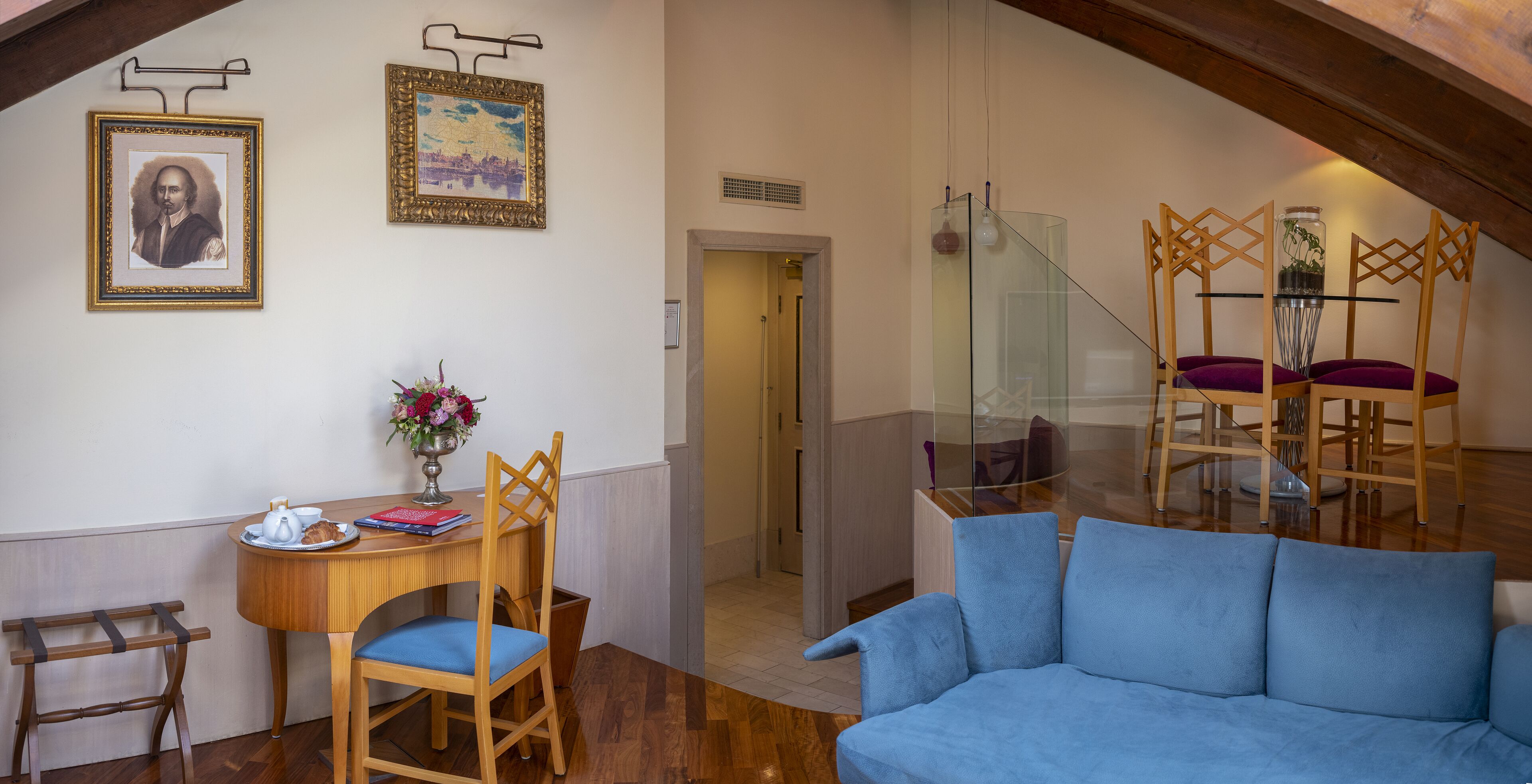 A cozy living room with a blue sofa, wooden chairs, a small round table with a vase of flowers, and framed pictures on the wall.