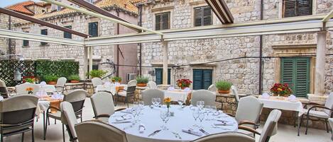 Un espace de restauration en plein air avec des tables rondes disposées pour deux personnes, entouré de bâtiments en pierre aux toits de tuiles.