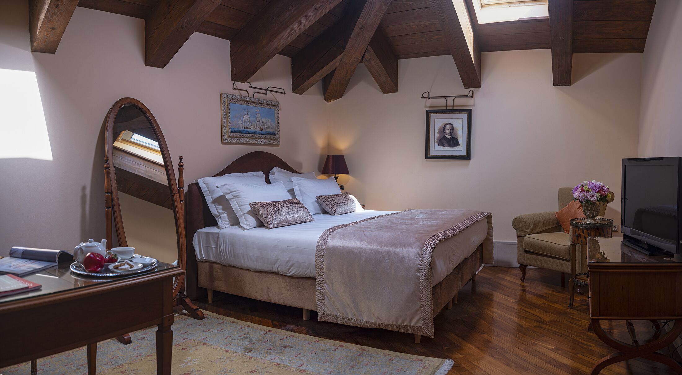 Ein Schlafzimmer mit einem großen Bett, einem Schreibtisch mit Spiegel, einem Sessel, einem Fernseher und einem Gemälde an der Wand.